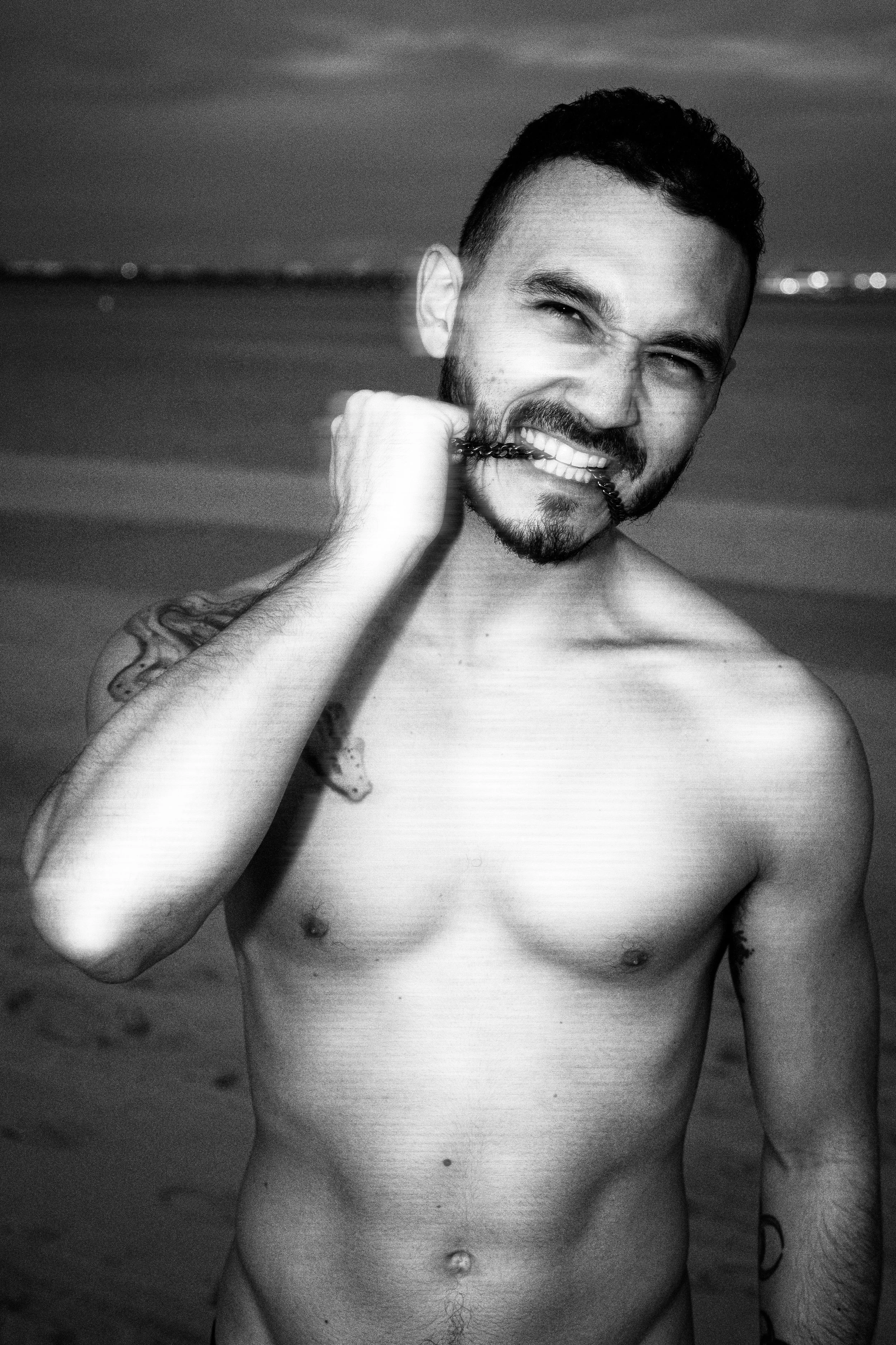 A shirtless man with tattoos and a beard biting a chain in his mouth on the beach during twilight, smiling at the camera.