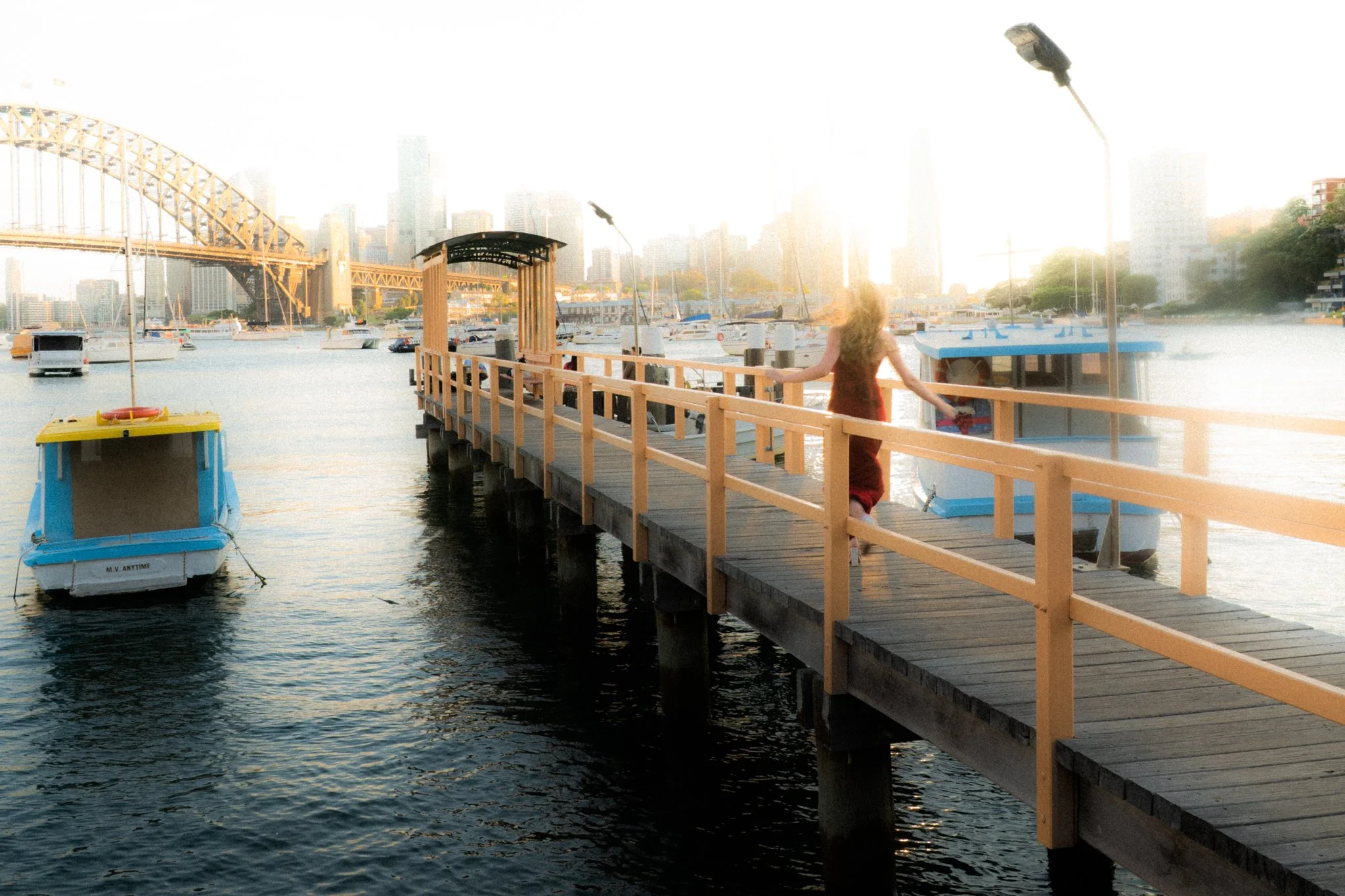 A woman walking on a wooden pier at a marina with boats, water, and city skyline in the background during sunset.