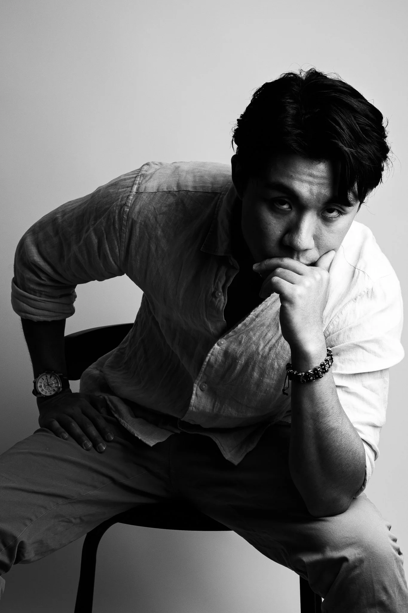 Black and white portrait of a man sitting on a chair, leaning forward with his hand near his face, dressed in a button-up shirt and pants, wearing a watch and bracelet.