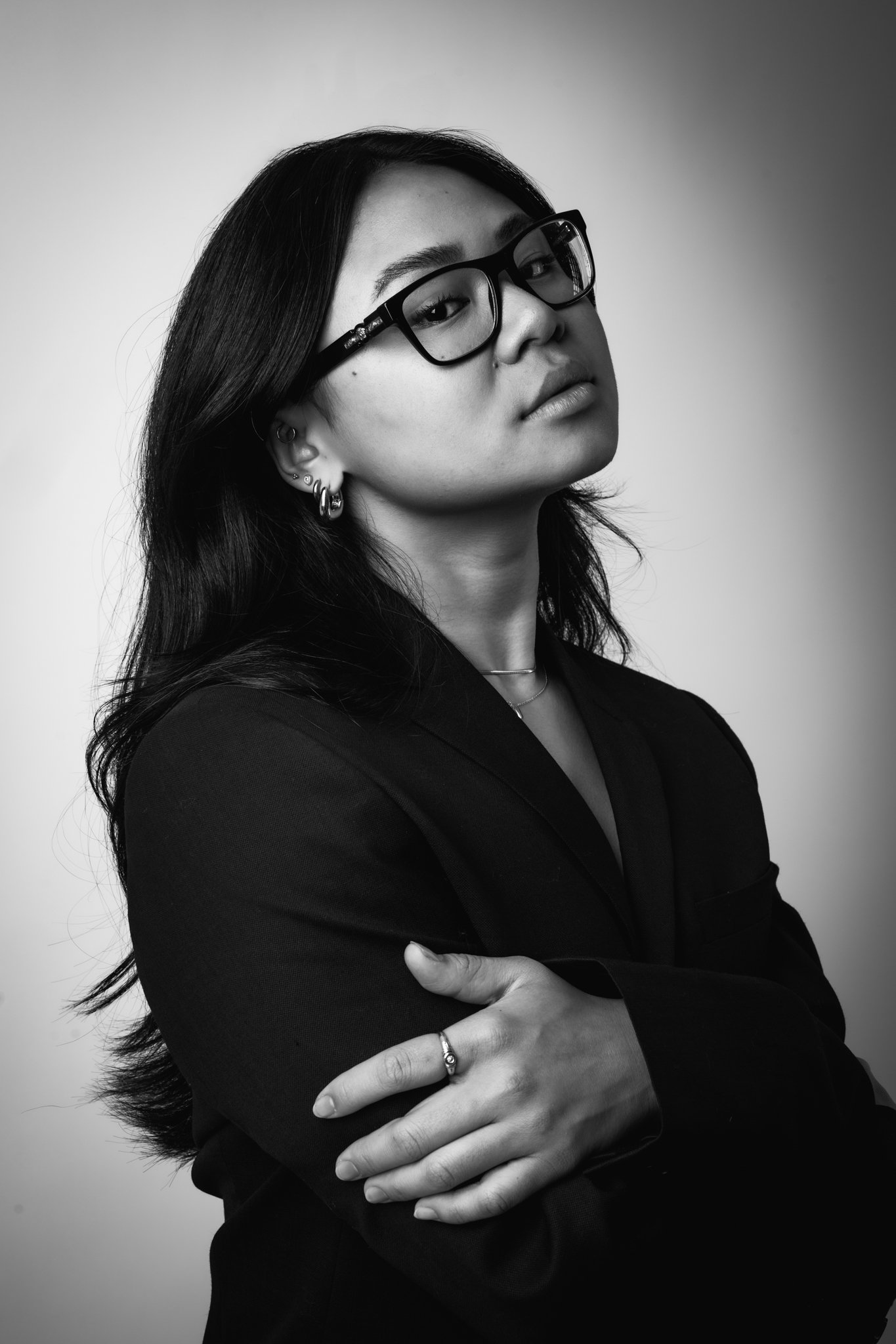 Black and white portrait of a woman with glasses, layered earrings, a necklace, and a blazer, crossing her arms and looking confidently at the camera.