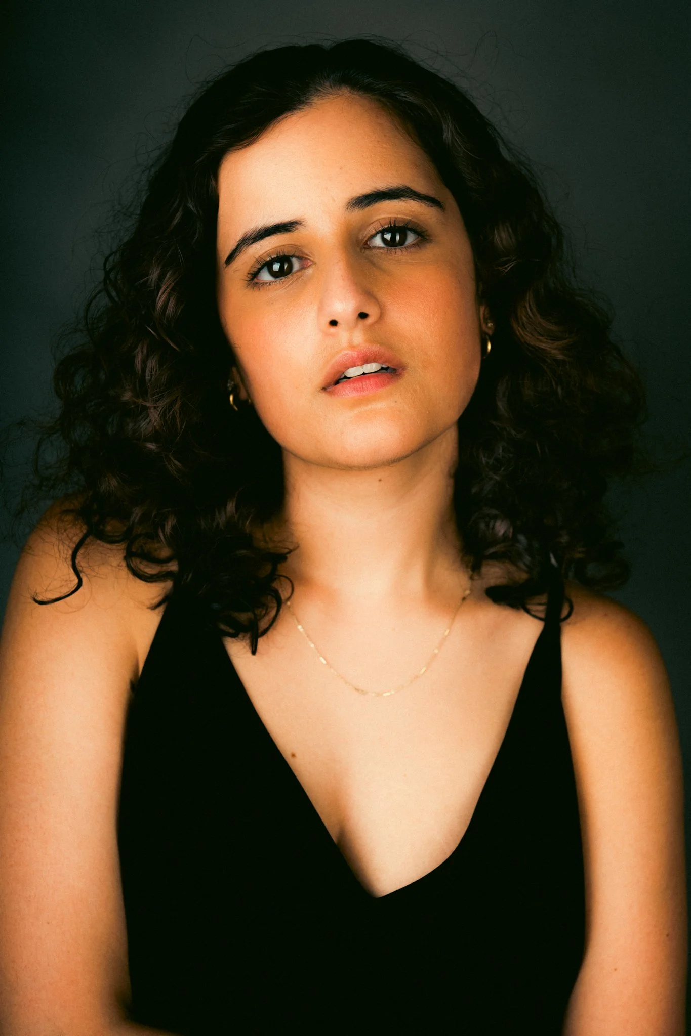 A woman with curly dark hair wearing a black sleeveless top, gold hoop earrings, and a delicate chain necklace, looking directly at the camera against a dark background.