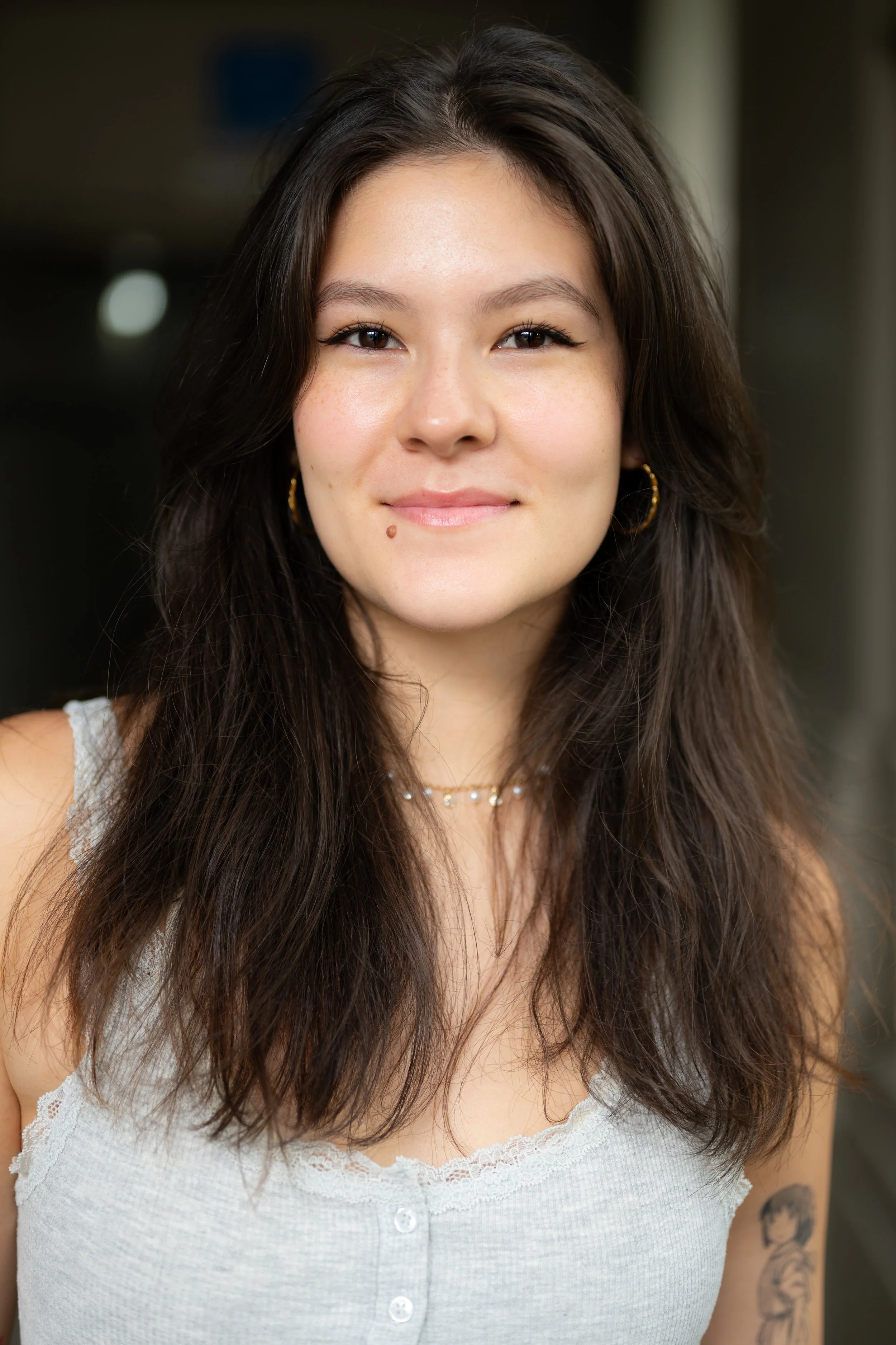 Close-up portrait of a young woman with long brown hair, smiling softly, wearing a white tank top with lace trim, gold hoop earrings, layered necklaces, and a tattoo on her upper arm, indoors with blurred background.