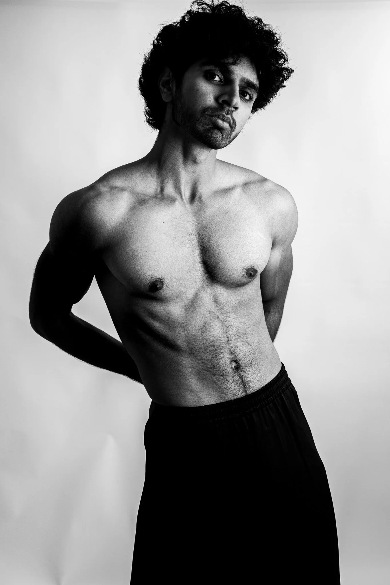 A black and white photo of a shirtless man with curly hair, standing against a plain background. He has a serious expression with slightly pursed lips, his head tilted, and his arms behind his back.