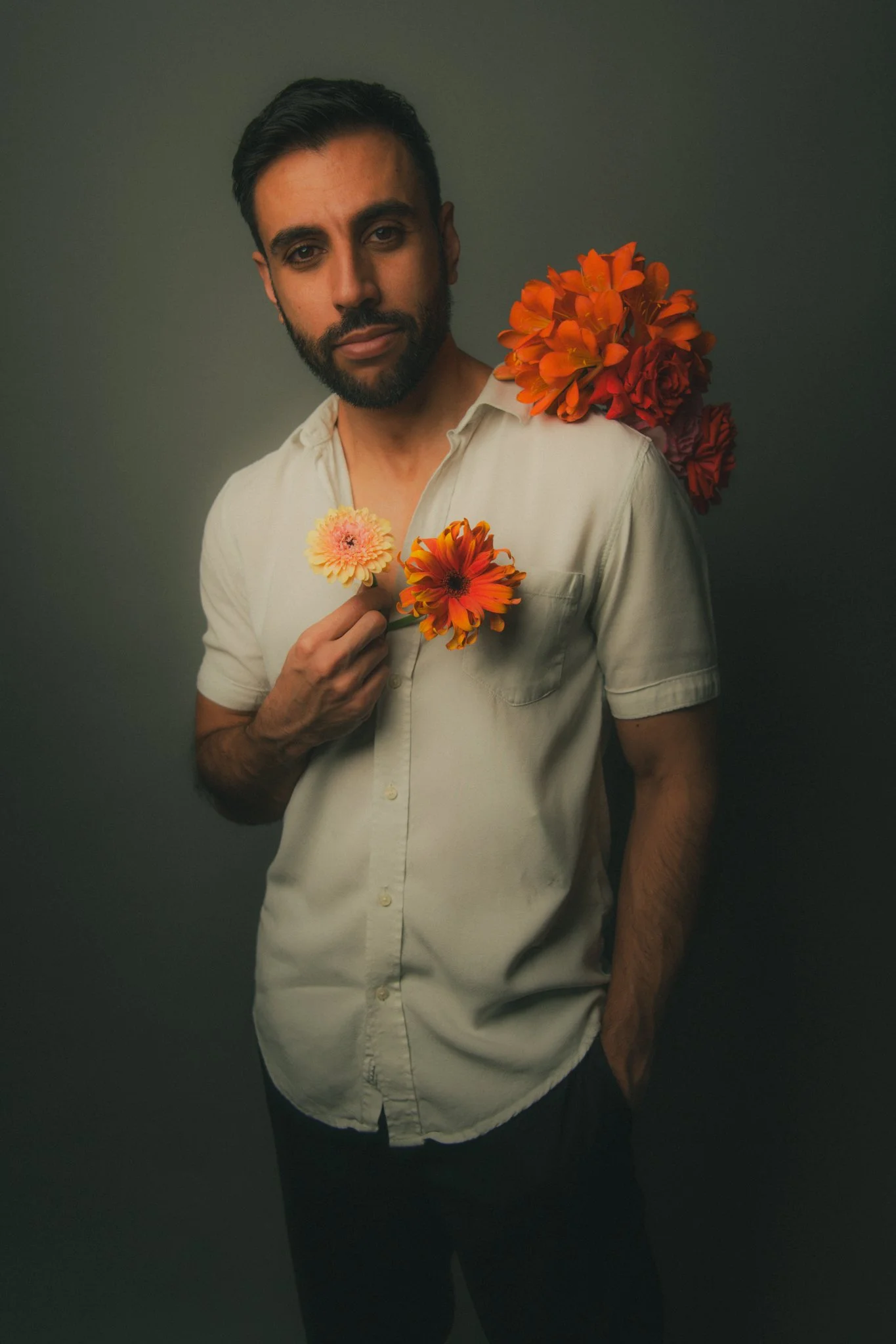 A man with short dark hair and a beard wearing a light-colored short-sleeve shirt, holding a pink flower, with orange and red flowers on his shoulder and back, against a dark background.