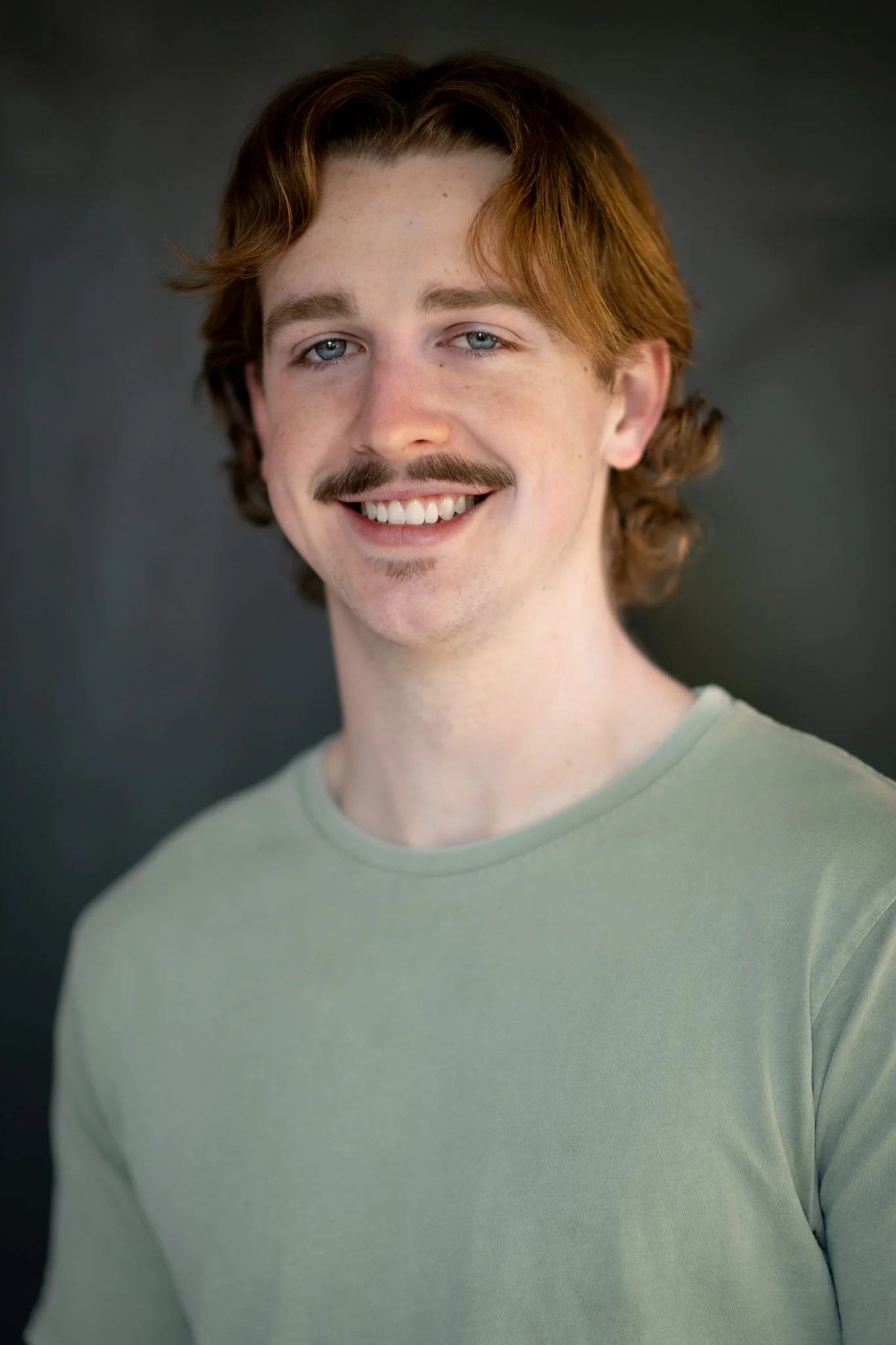 A young man with red hair and a mustache, smiling while wearing a light gray T-shirt, standing against a dark background.