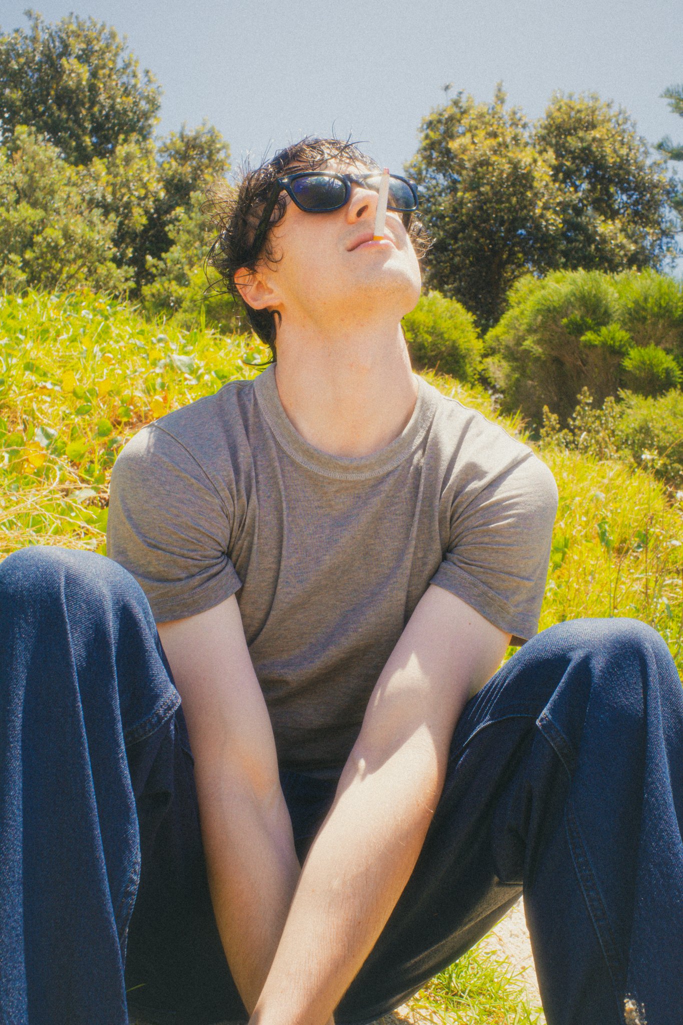 A young man with dark, curly hair and sunglasses sits outdoors on grass, leaning back with his head tilted upwards, holding a cigarette in his mouth, in a sunny park with trees in the background.