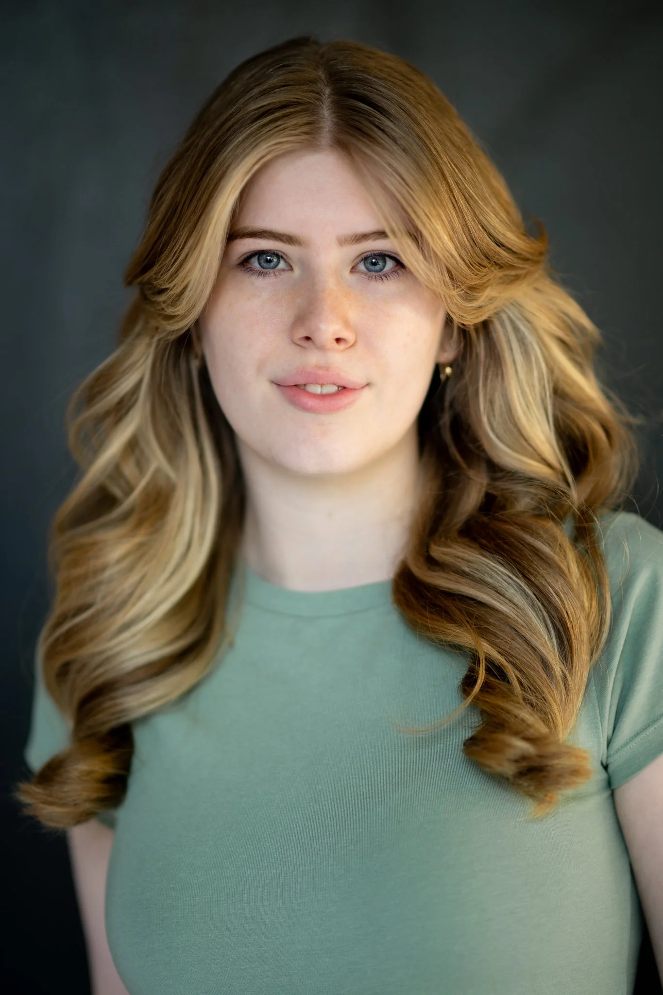 A portrait of a young woman with long, wavy, red hair, blue eyes, and fair skin, wearing a light green T-shirt against a dark background.