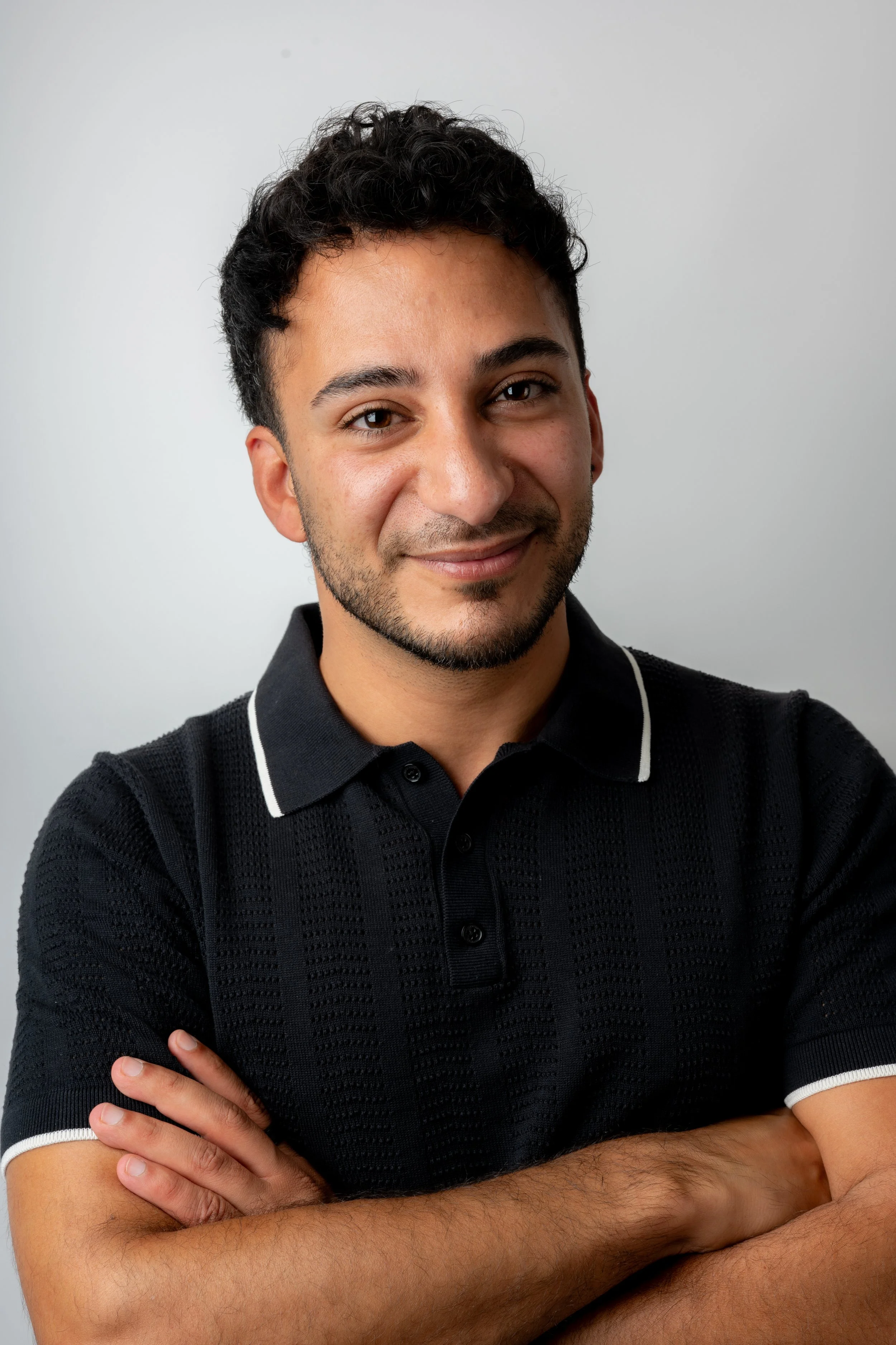 Portrait of a young man with short curly black hair, light brown skin, wearing a black polo shirt with white trim, smiling with arms crossed in front of a plain light gray background.