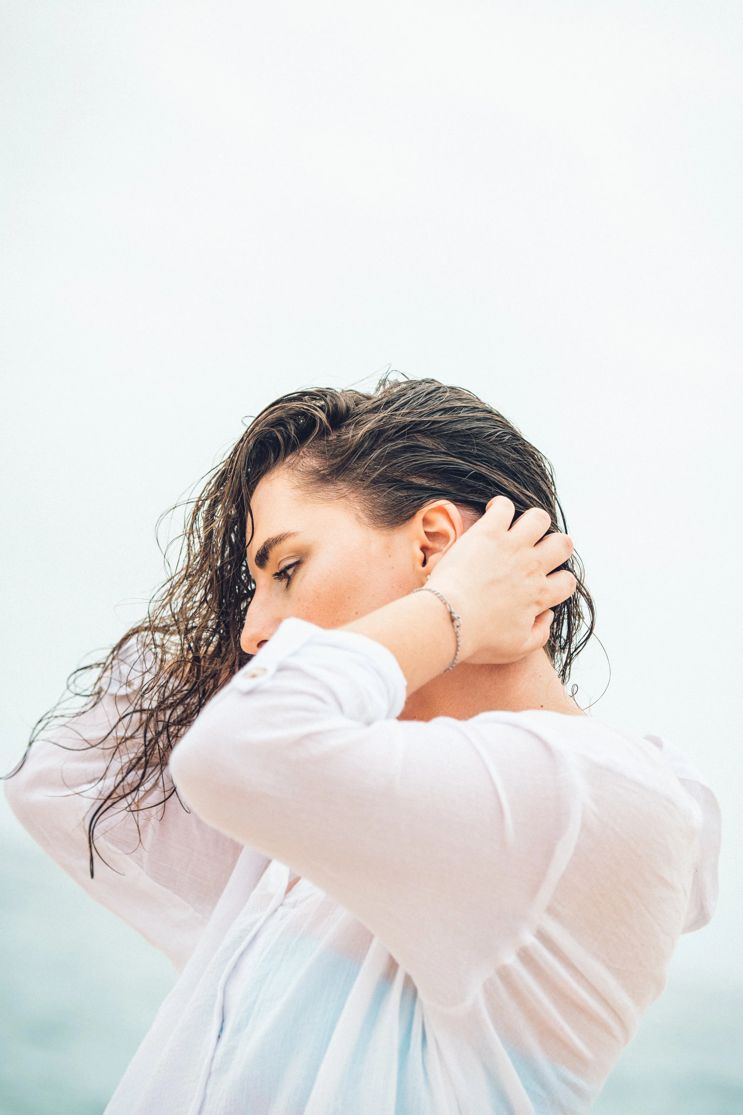 A woman with wet hair, wearing a white shirt, is outdoors, touching her neck with both hands, with a calm and pensive expression.