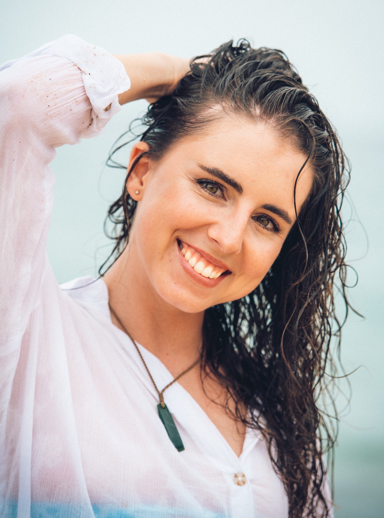 A woman with wet, curly hair smiling and touching her head outdoors near water.