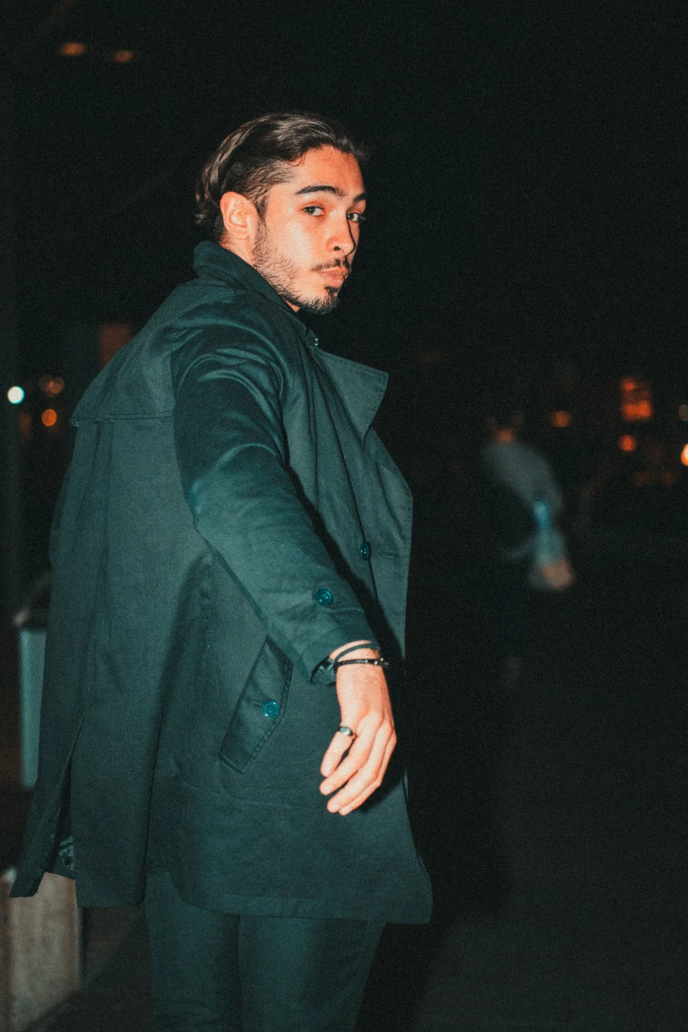 A young man with dark hair and a beard, dressed in a black coat, standing outdoors at night.