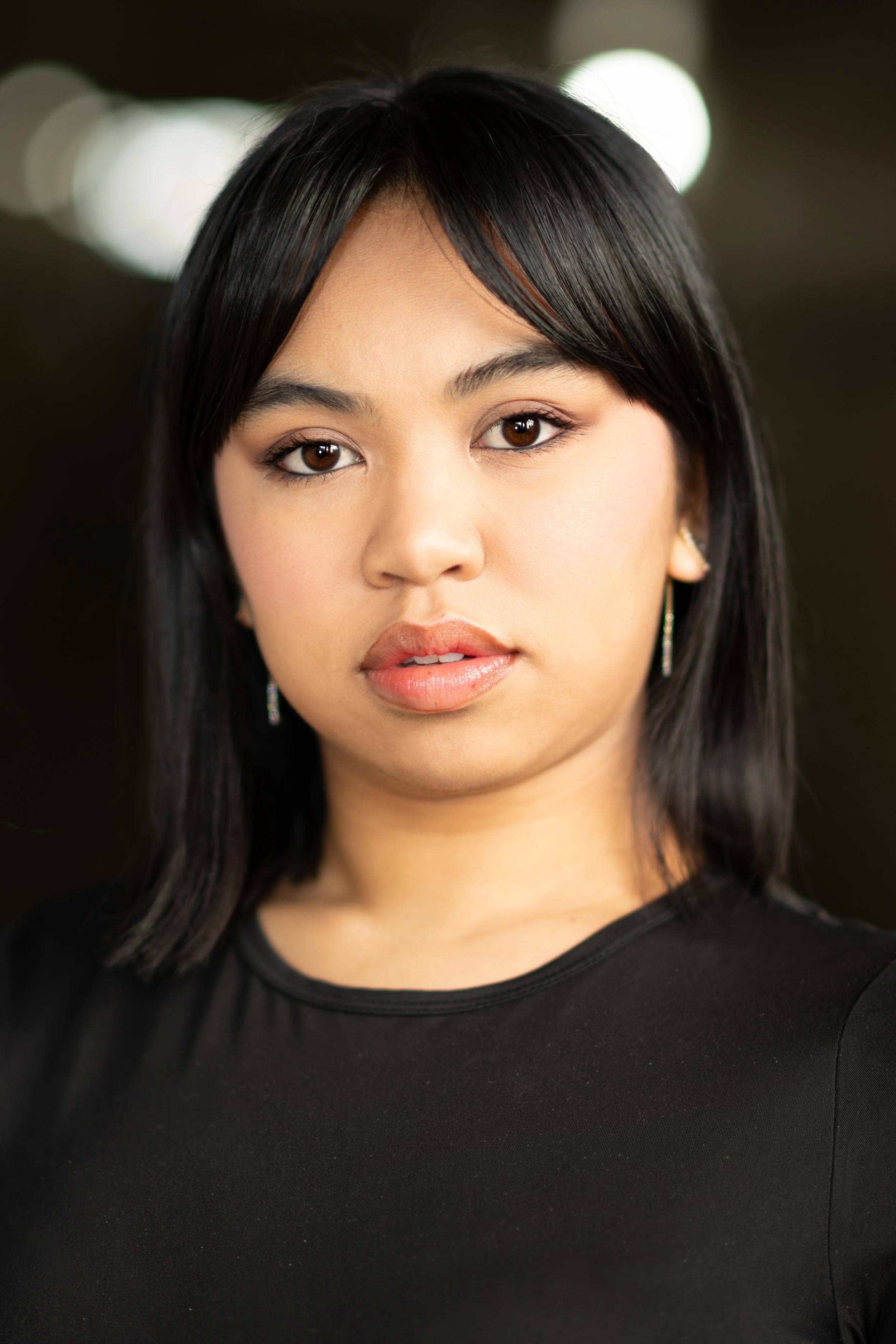 Close-up of a young woman with straight black hair, wearing a black top, earrings, and subtle makeup, looking directly at the camera.
