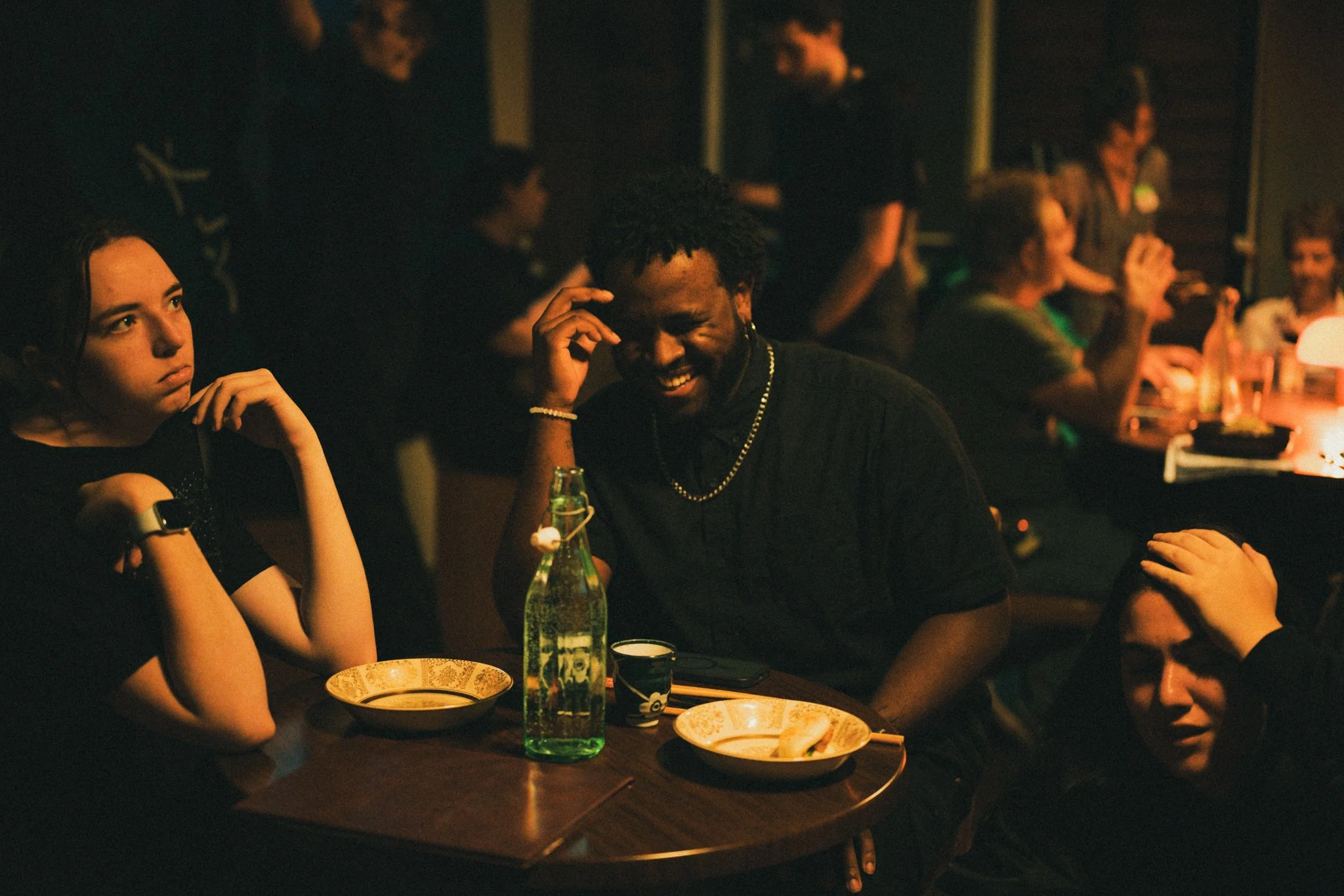 People sitting at a restaurant table. The focus is on a man laughing, surrounded by women. The scene is dimly lit, and there are bottles and dishes on the table.