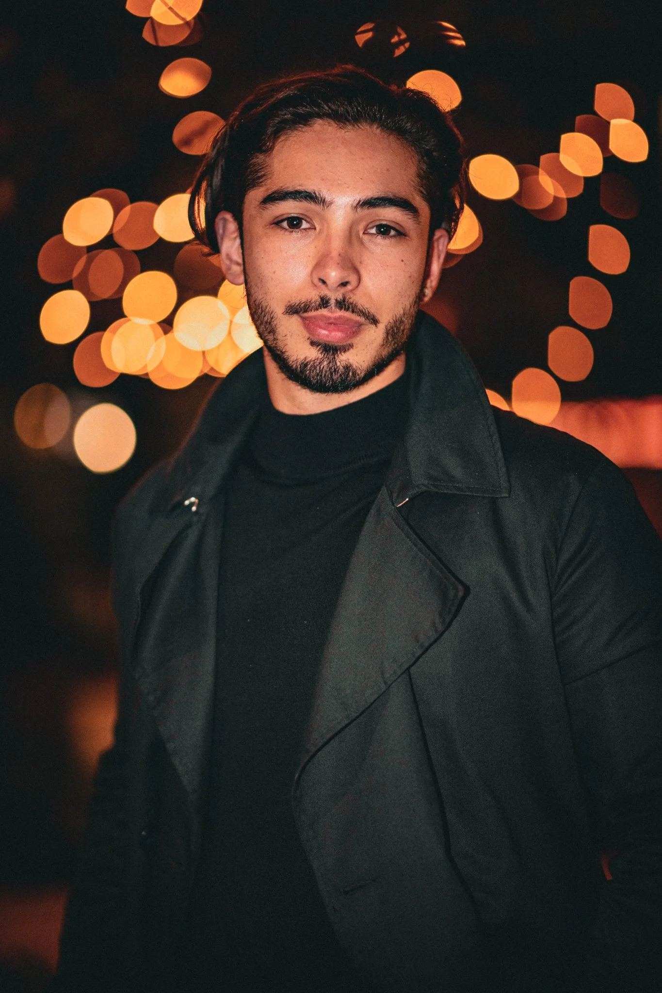 A young man with dark hair and a beard posing outdoors at night, with blurred orange and yellow lights in the background.
