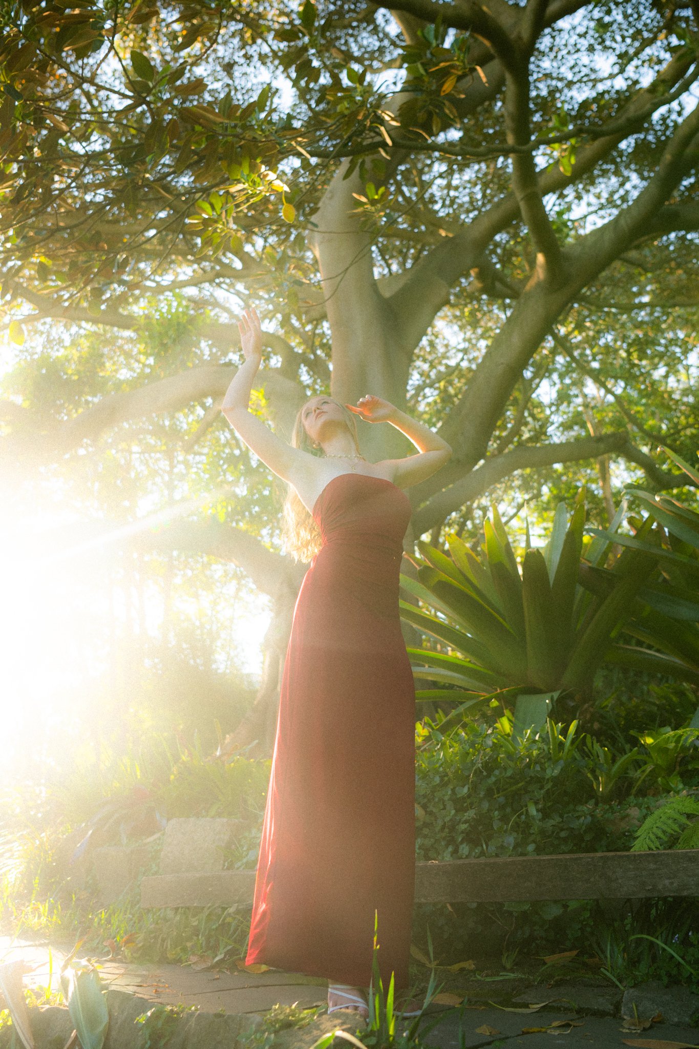 A woman in a strapless maroon dress standing outdoors among greenery, partially illuminated by sunlight, with arms raised and head tilted back.