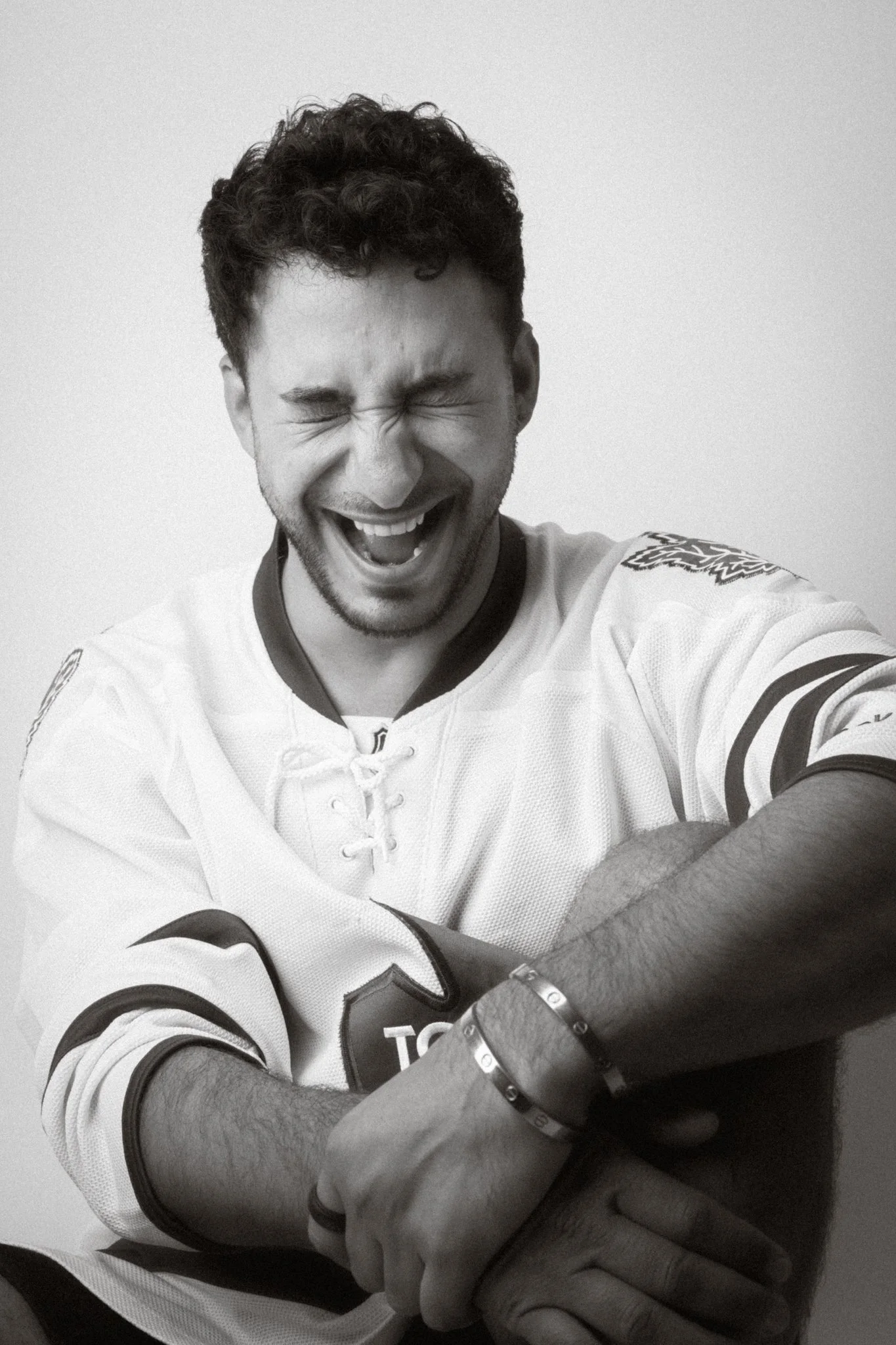 A man with curly hair wearing a hockey jersey, smiling with eyes closed and arms crossed, posing against a plain background.