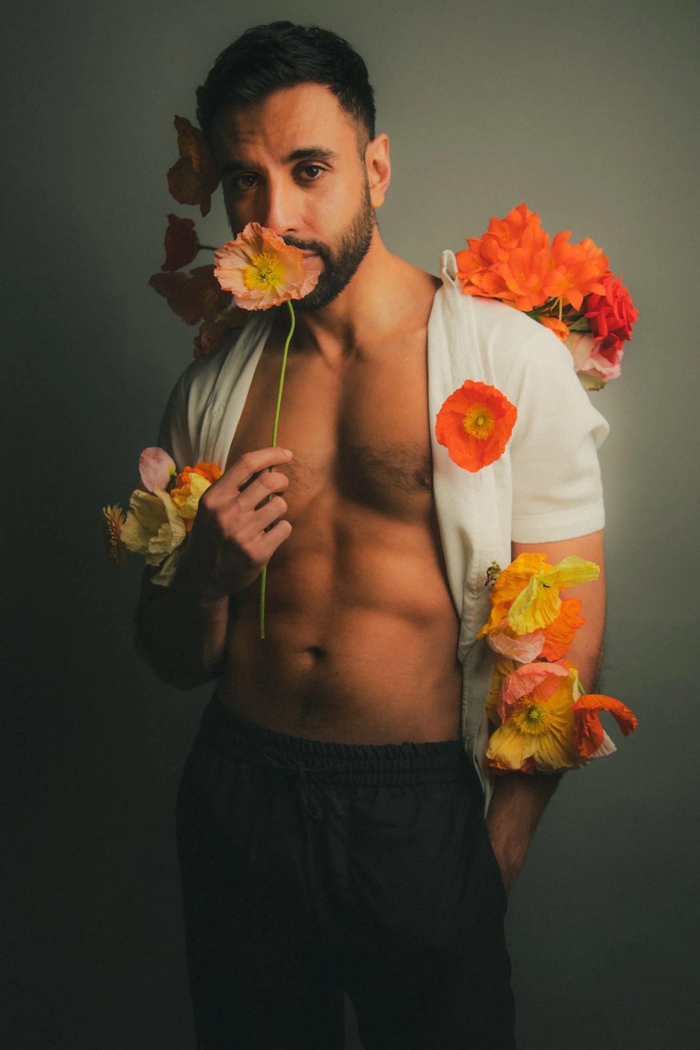 A shirtless man with dark hair and a beard holding a pink flower near his face, surrounded by colorful flowers on his shoulder, arm, and jacket, posing against a plain gray background.