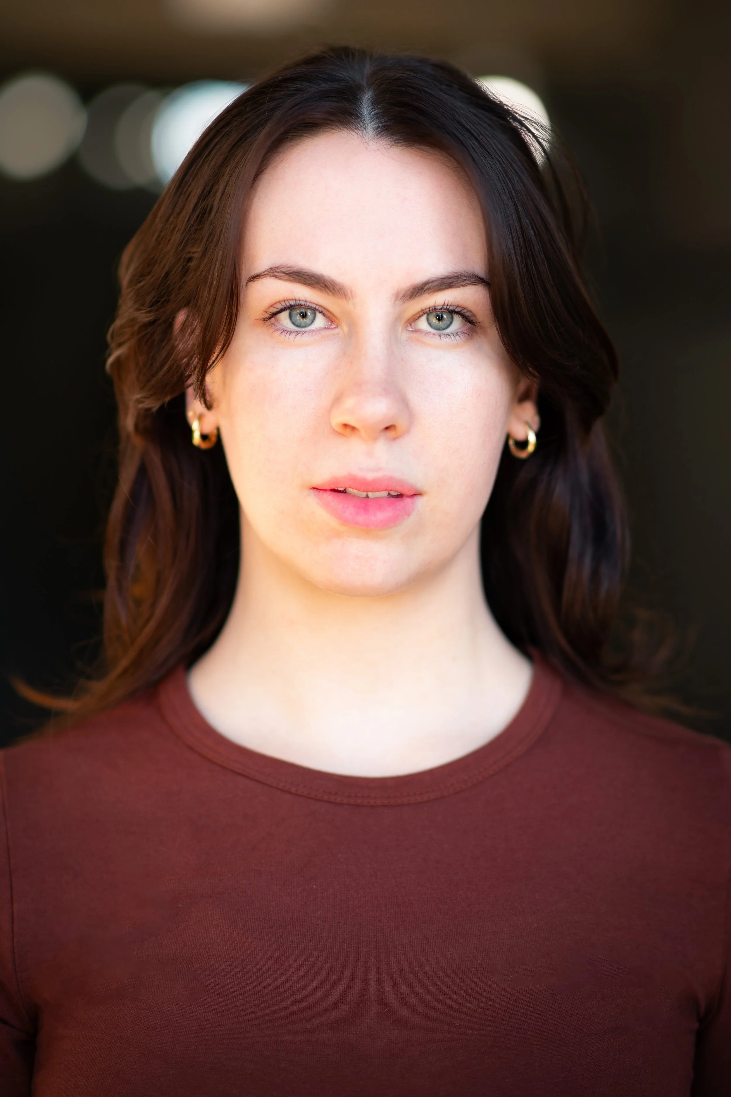 Close-up portrait of a young woman with blue eyes and dark brown hair, wearing gold hoop earrings and a maroon top, against a blurred dark background.