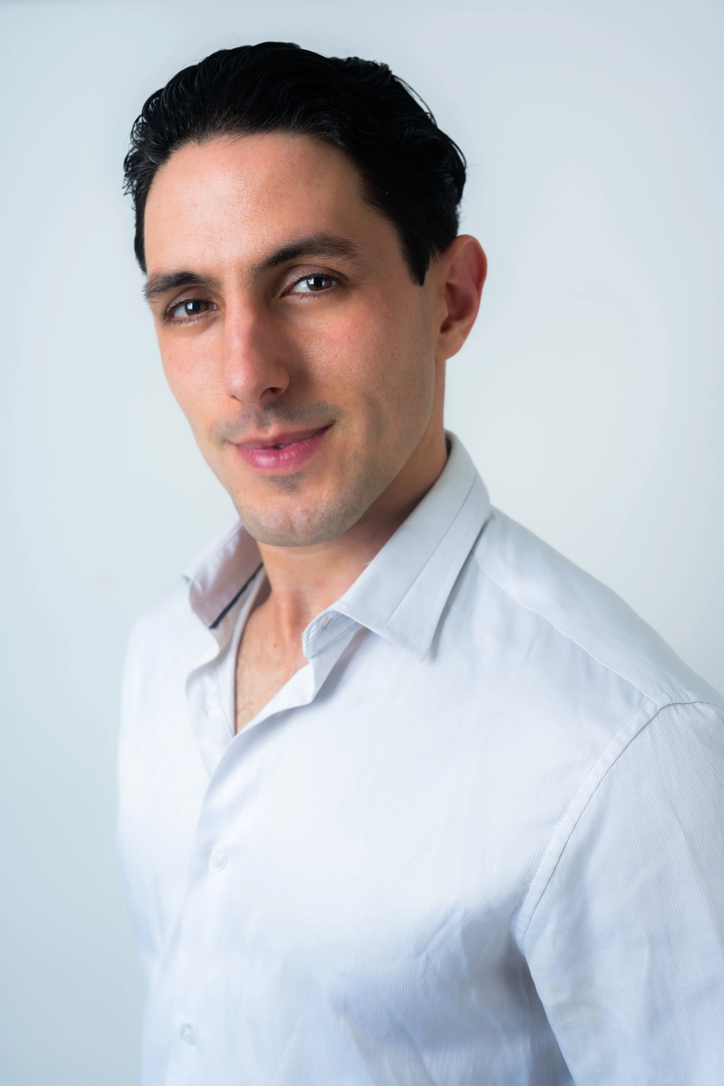 A young man with dark hair styled back, wearing a white dress shirt, smiling slightly, with a light gray background.