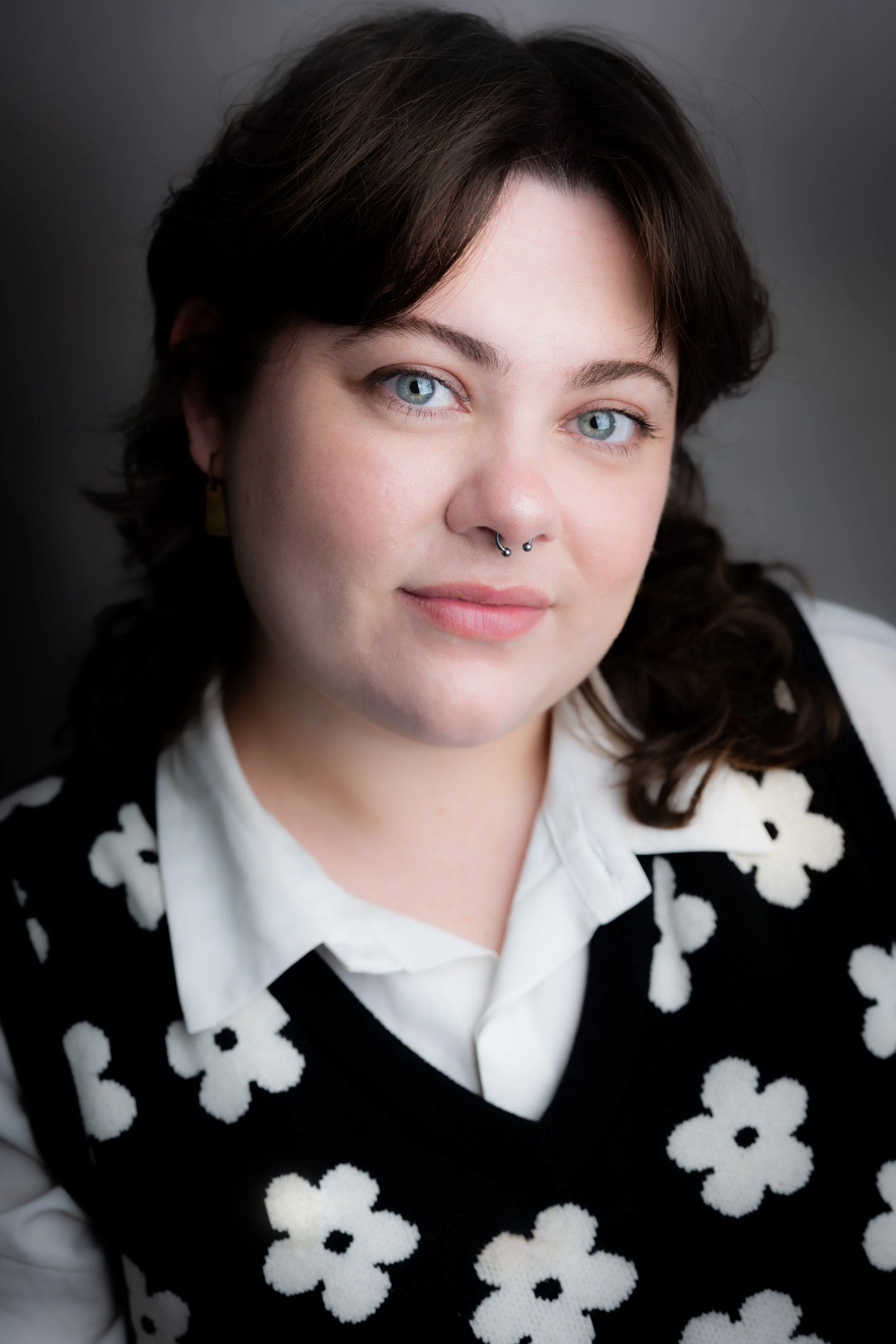 Close-up portrait of a woman with blue eyes, dark brown hair, wearing a black vest with white flower patterns over a white collared shirt, and a septum piercing.