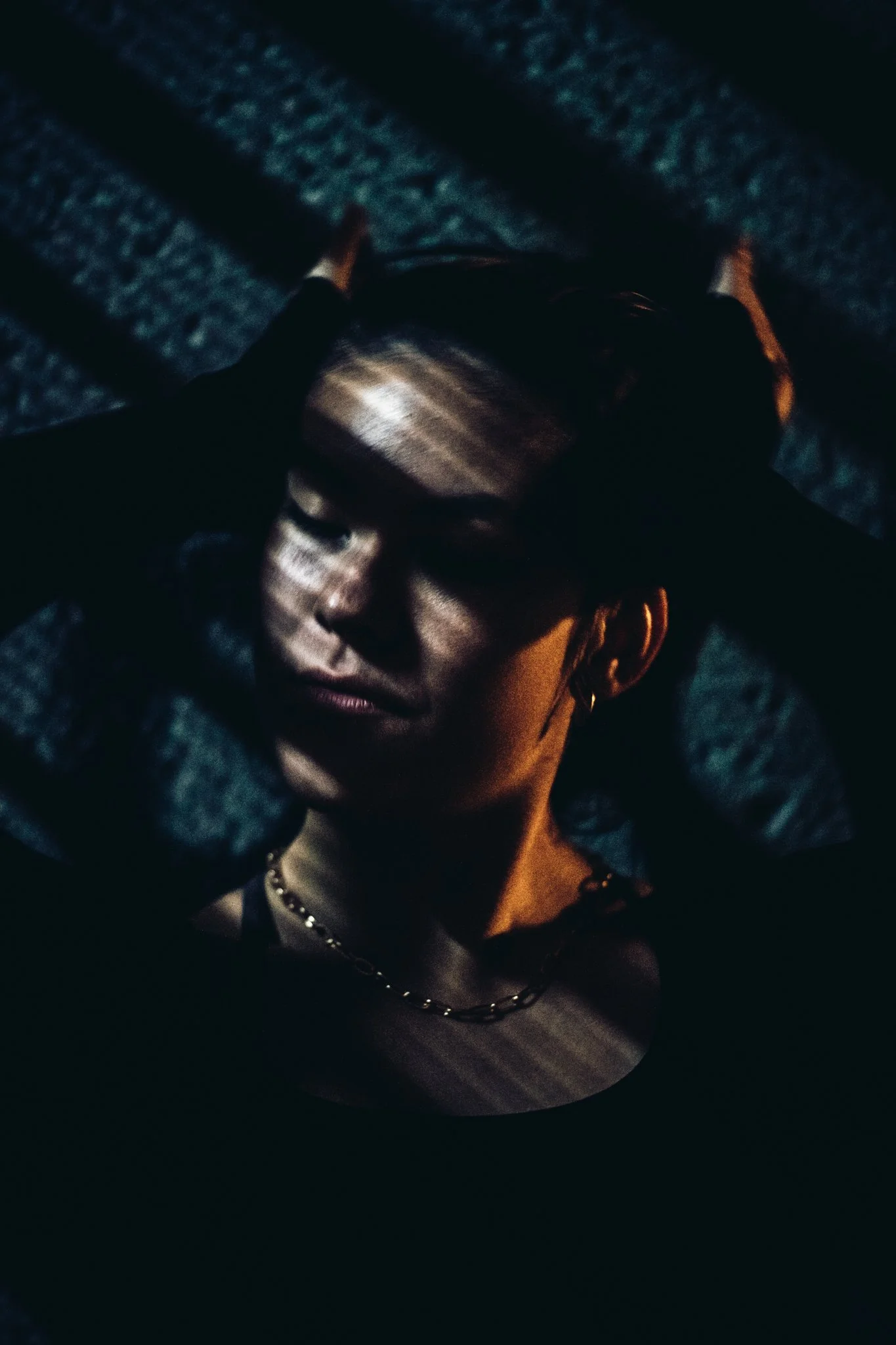 A woman with closed eyes and a peaceful expression, shadowed with light and dark stripes, lying on a textured surface with her arms raised above her head, wearing a black top, a chain necklace, and hoop earrings.