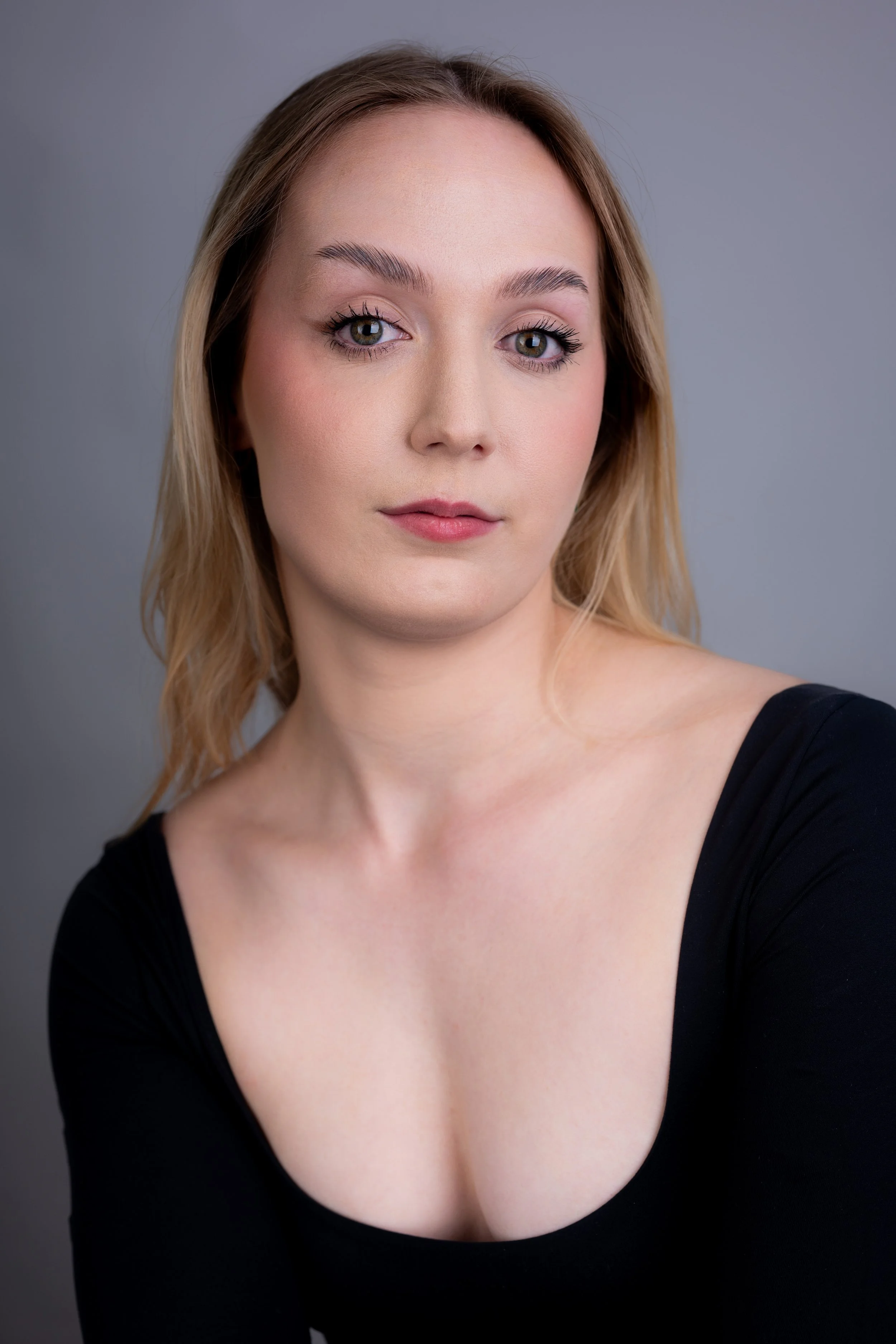 Portrait of a young woman with blonde hair wearing a black top, looking at the camera against a gray background.