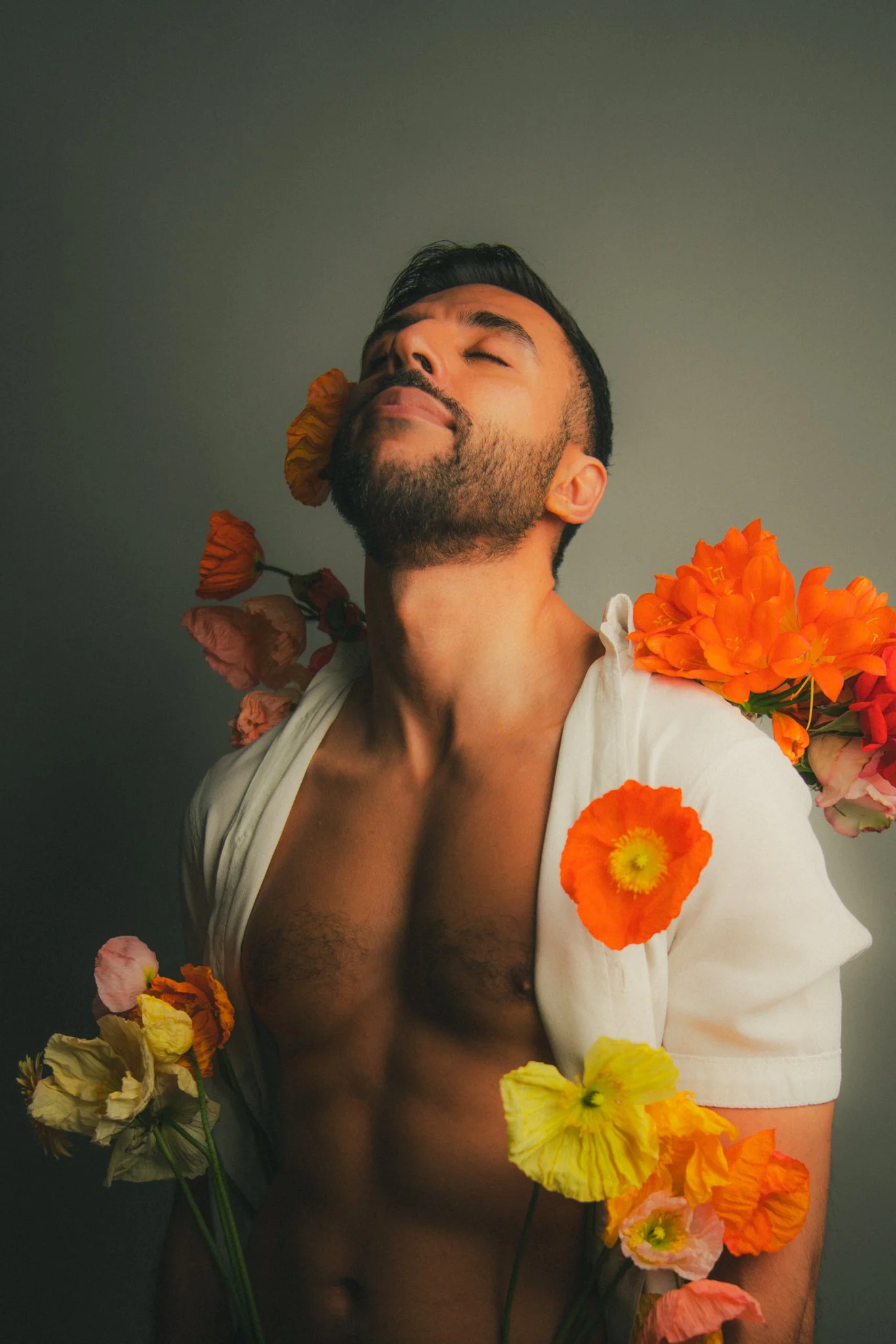 A shirtless man with closed eyes and a content expression, surrounded by and wearing colorful flowers on his shoulders and chest, against a plain background.