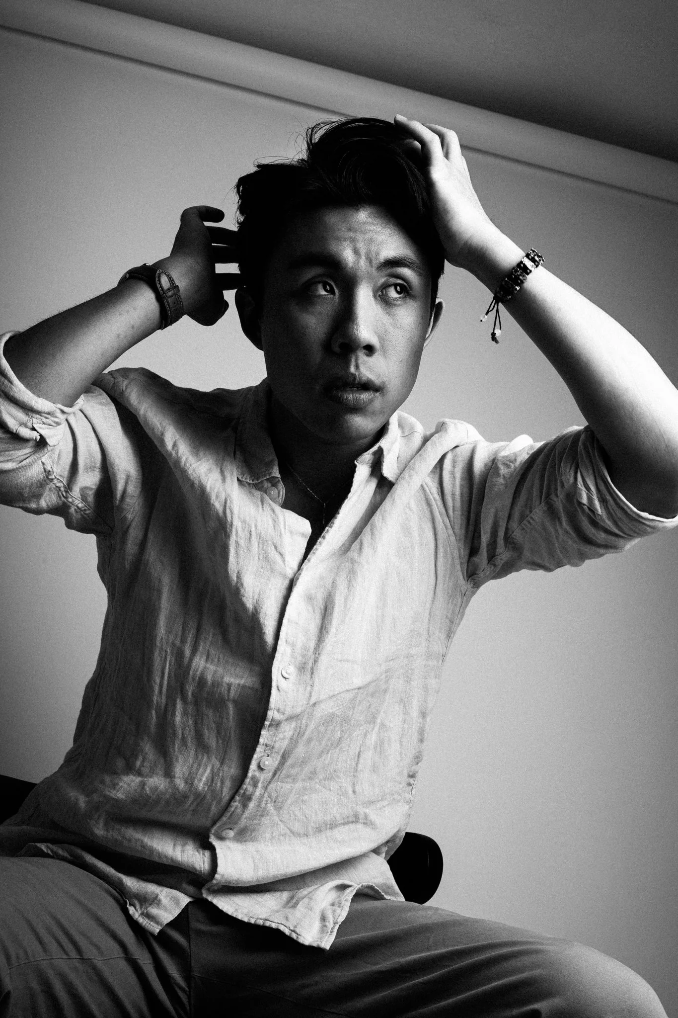 A young man with dark hair, wearing a light-colored, button-up shirt and multiple bracelets, sitting on a chair, touching his hair in a contemplative pose in a black-and-white photo.