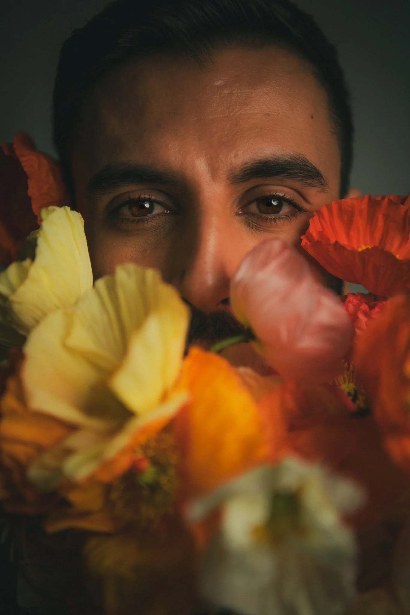 Close-up of a man's face peeking through colorful flowers, with only his eyes and part of his forehead visible.