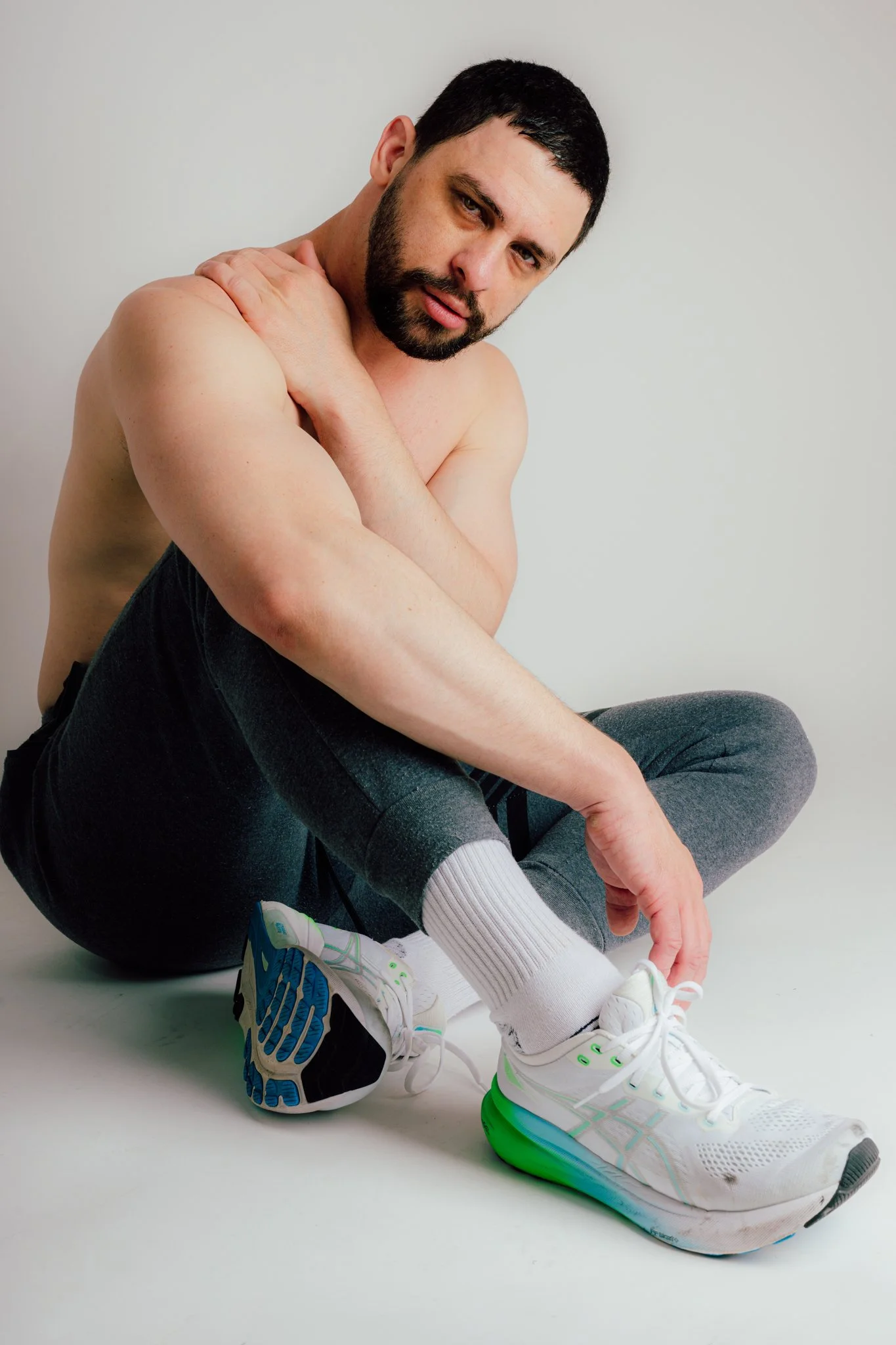 A shirtless man with dark hair and a beard sitting on the floor, adjusting his running shoe.