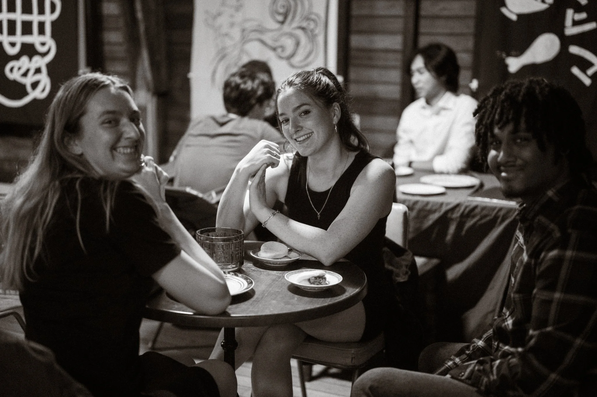 Four friends sitting at a small round table in a restaurant, smiling and enjoying their time. Two women in the foreground and two people in the background, with plates and a drink on the table, in a cozy, dimly lit setting with wooden walls and paint
