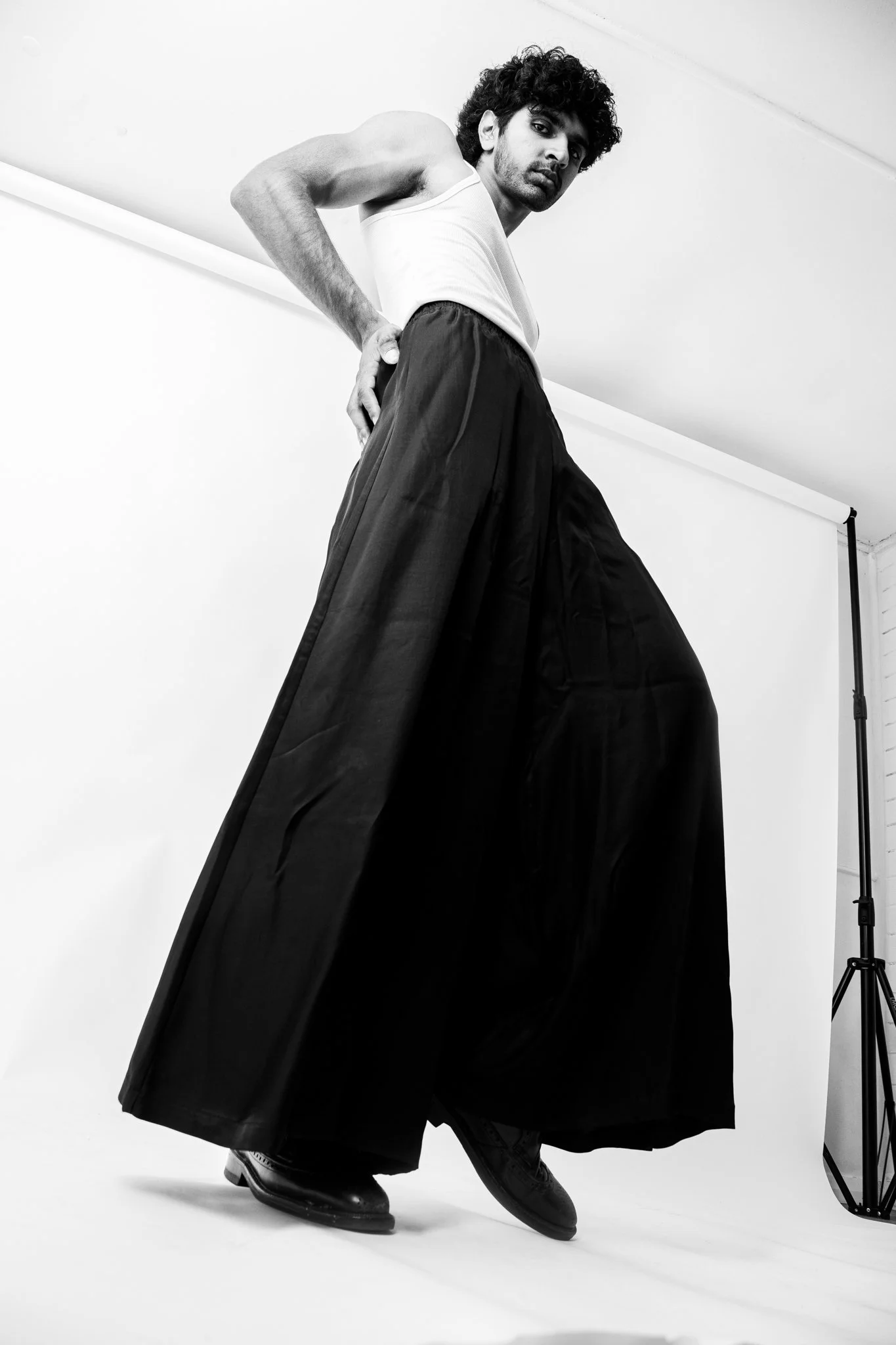 A man with curly hair and a beard poses in a studio with a white backdrop, wearing a white sleeveless shirt, wide black pants, and black shoes.