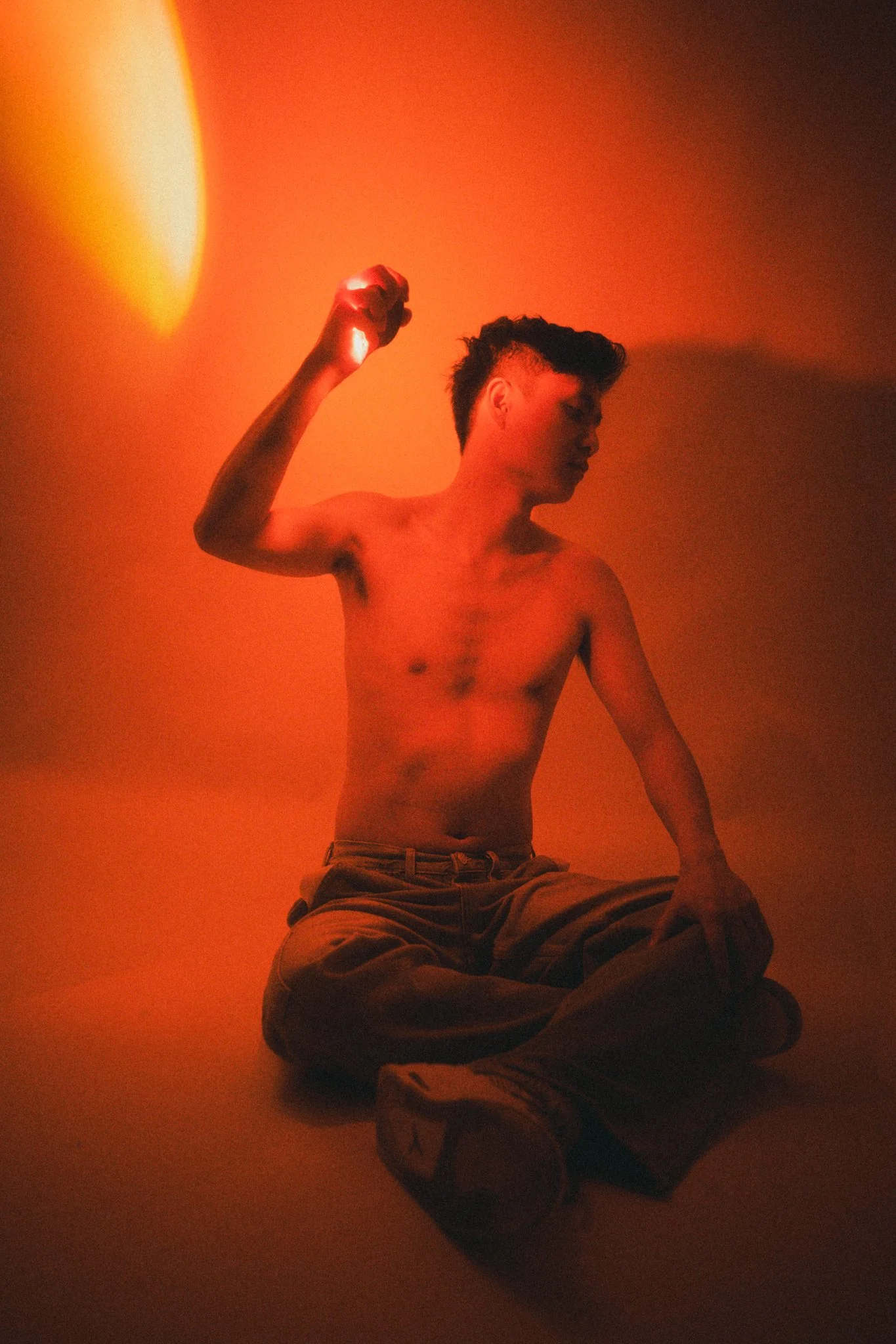 A shirtless young man sitting cross-legged on the floor in front of an orange wall, raising his right arm with a clenched fist, illuminated by a warm light source.