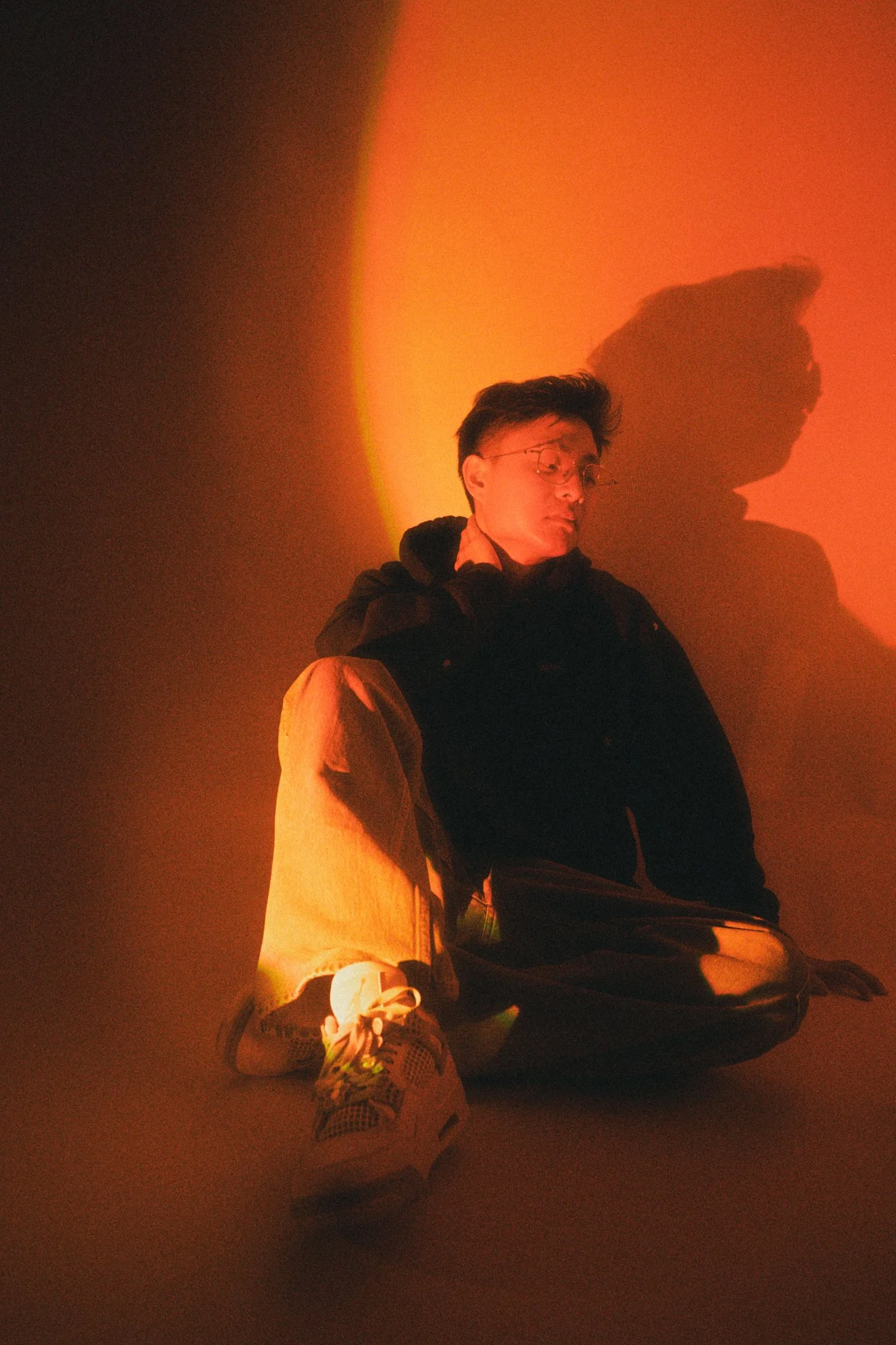 A person sitting on the floor with one knee up, illuminated by orange lighting, with their hand on their neck. The shadows and warm tones create a dramatic atmosphere.