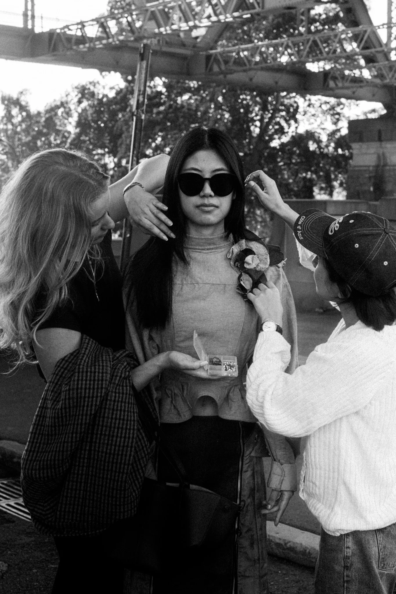 A woman with long dark hair wearing sunglasses is being styled or prepared by two assistants, one holding makeup or jewelry, at an outdoor location under a bridge or structure.