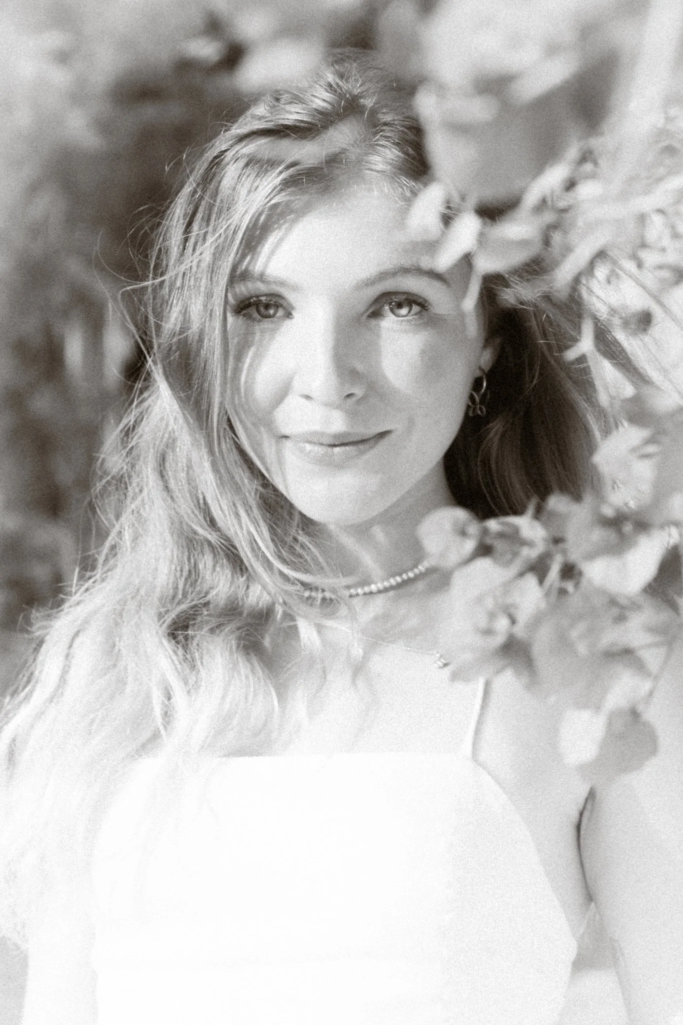 A woman with long wavy hair smiling outdoors, partly obscured by leaves.
