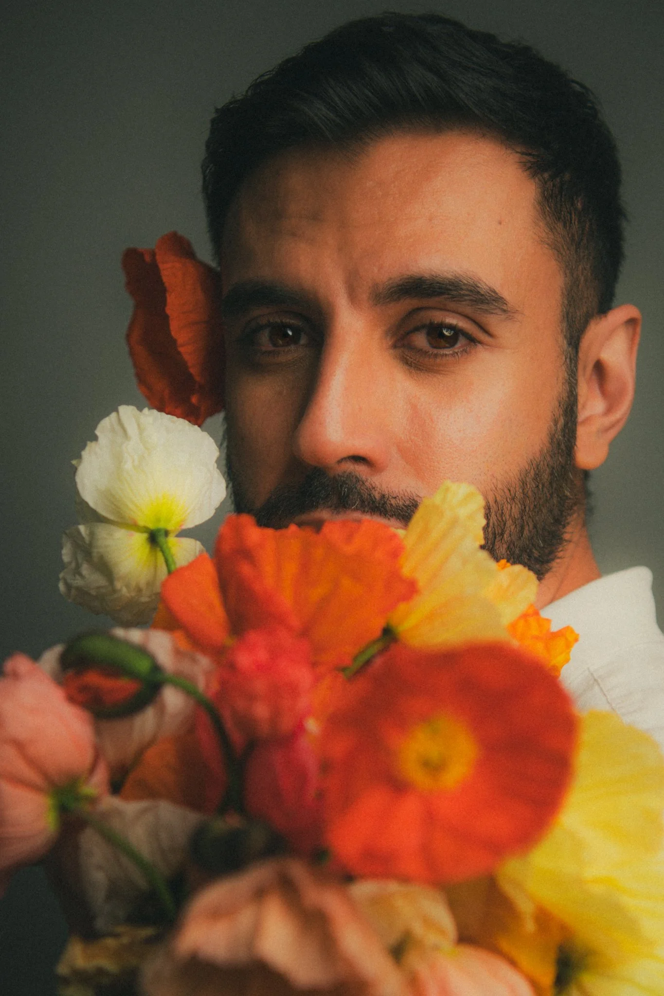 Close-up portrait of a man with dark hair and beard surrounded by colorful flowers, looking directly at the camera.