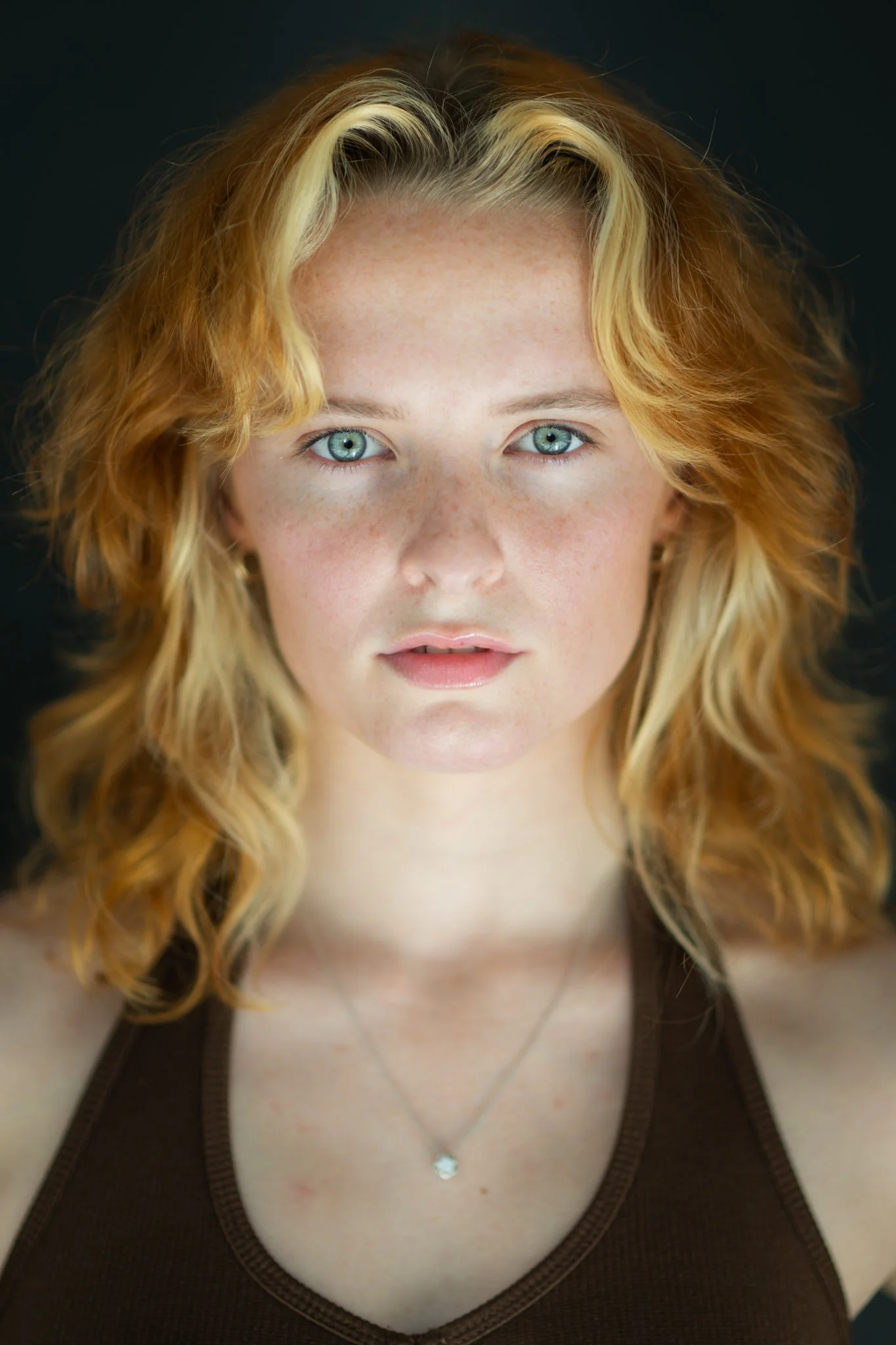 Close-up portrait of a woman with curly blonde hair, blue eyes, and freckles, wearing a brown sleeveless top and a silver necklace, against a dark background.