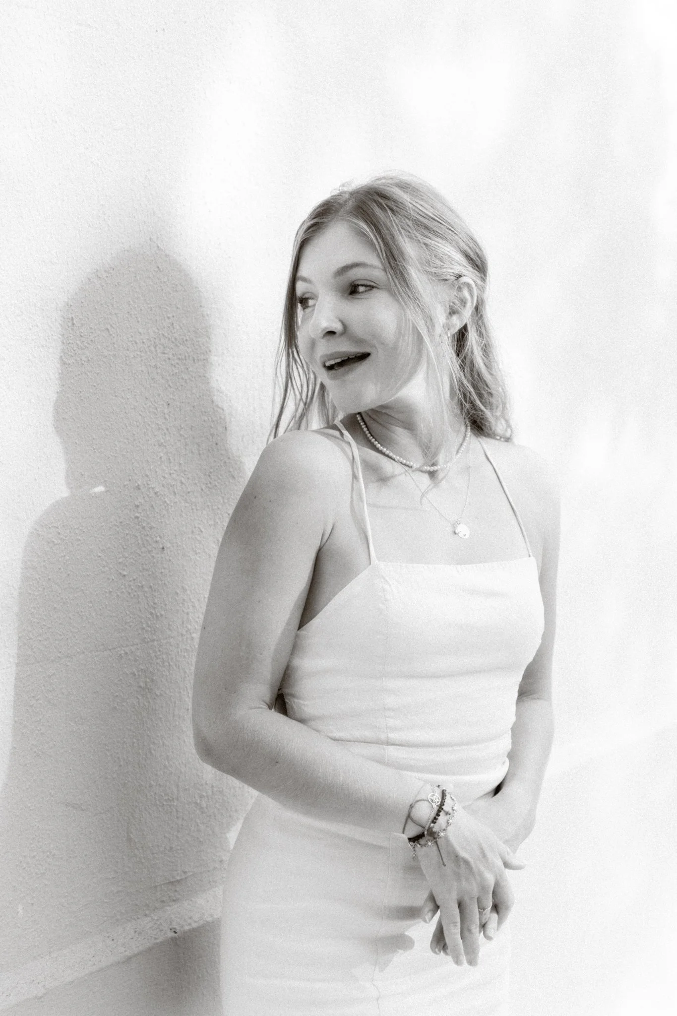 A woman with long hair in a white spaghetti strap dress standing against a textured wall, looking to her left with a smile.