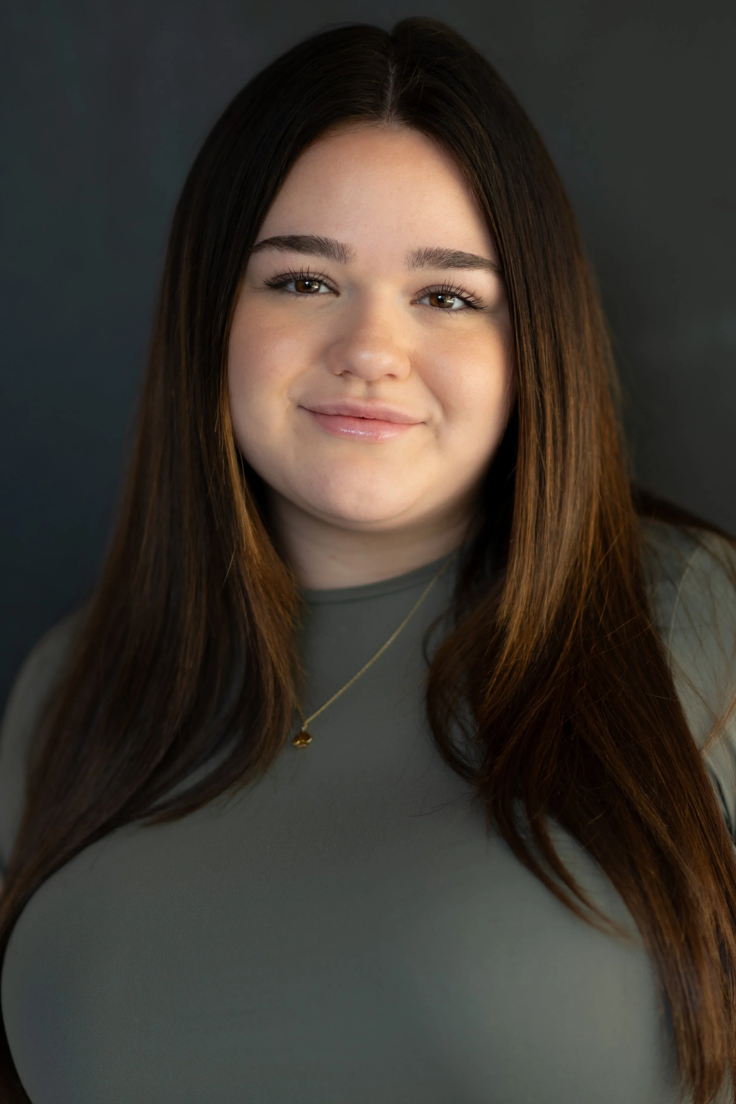 Close-up portrait of a young woman with long brown hair, smiling softly, wearing a gray top and a gold necklace, against a dark background.