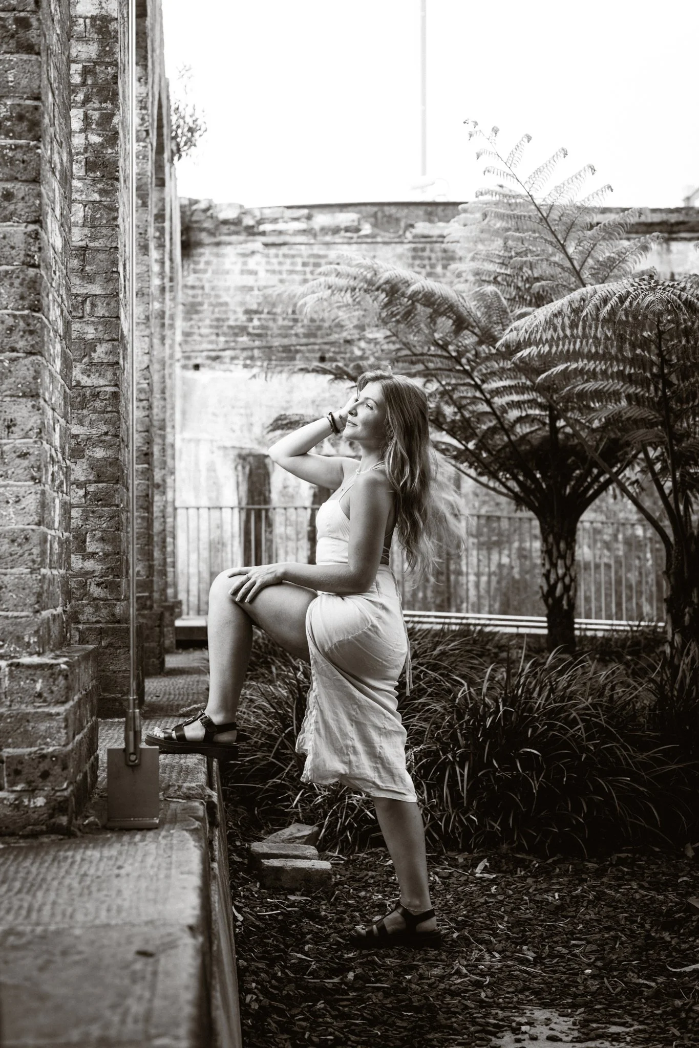 A woman in a dress standing outside on a garden step with one foot on the edge, touching her hair with one hand, and smiling with eyes closed, surrounded by plants and brick walls.