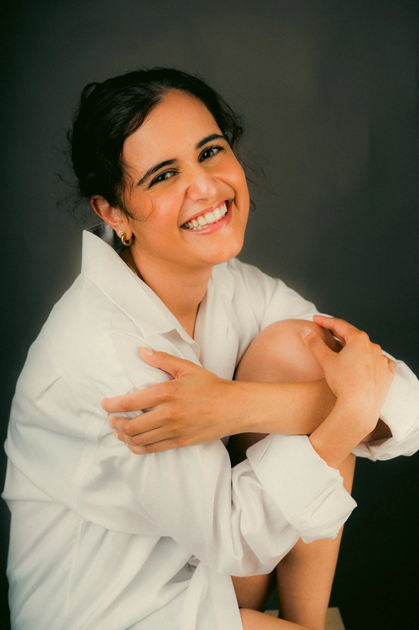 A woman with dark hair, wearing a white shirt, smiling, sitting with her arms around her knees against a dark background.