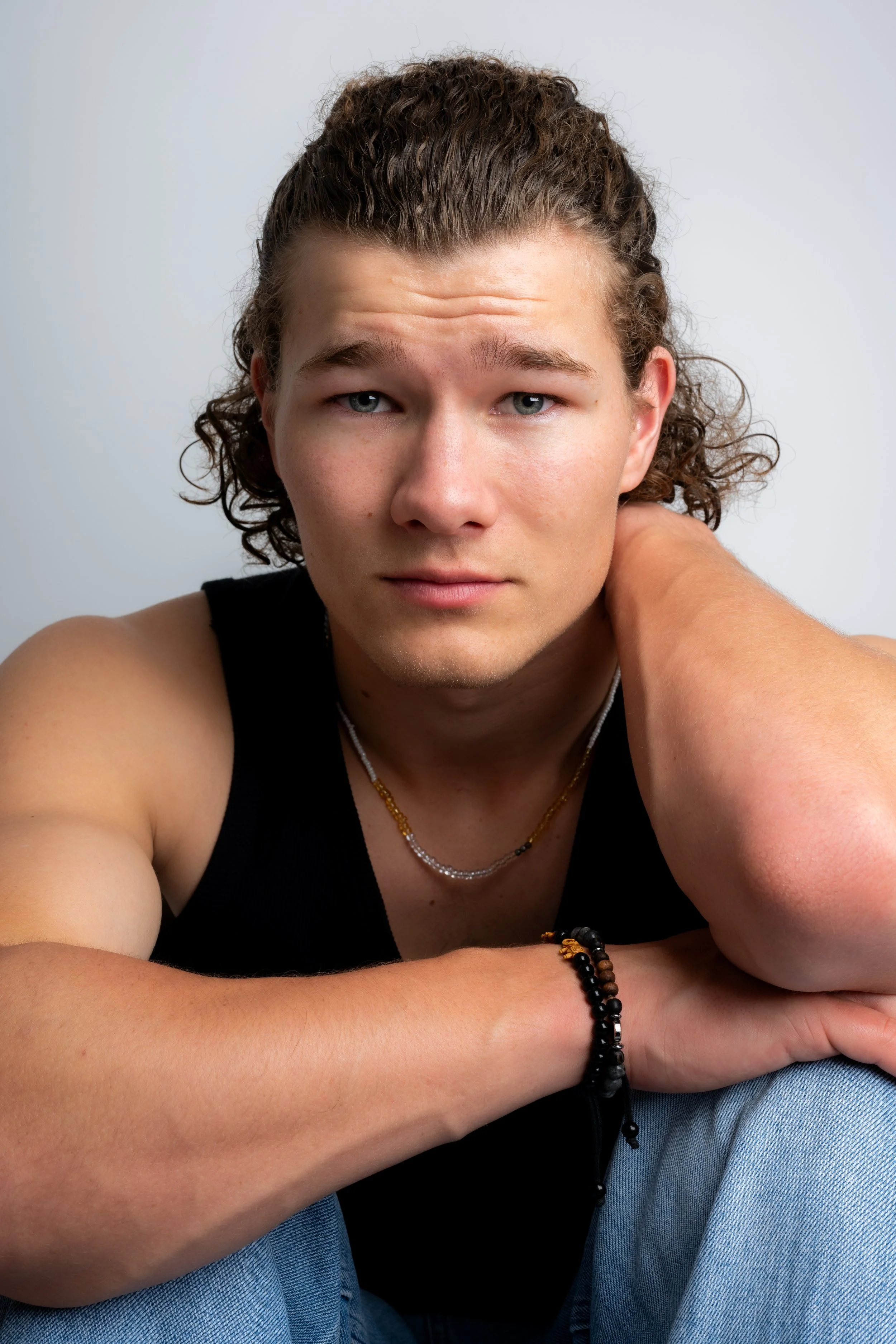 A young man with curly hair and a black sleeveless shirt, sitting against a plain background, resting his head on one hand, with a focused expression.