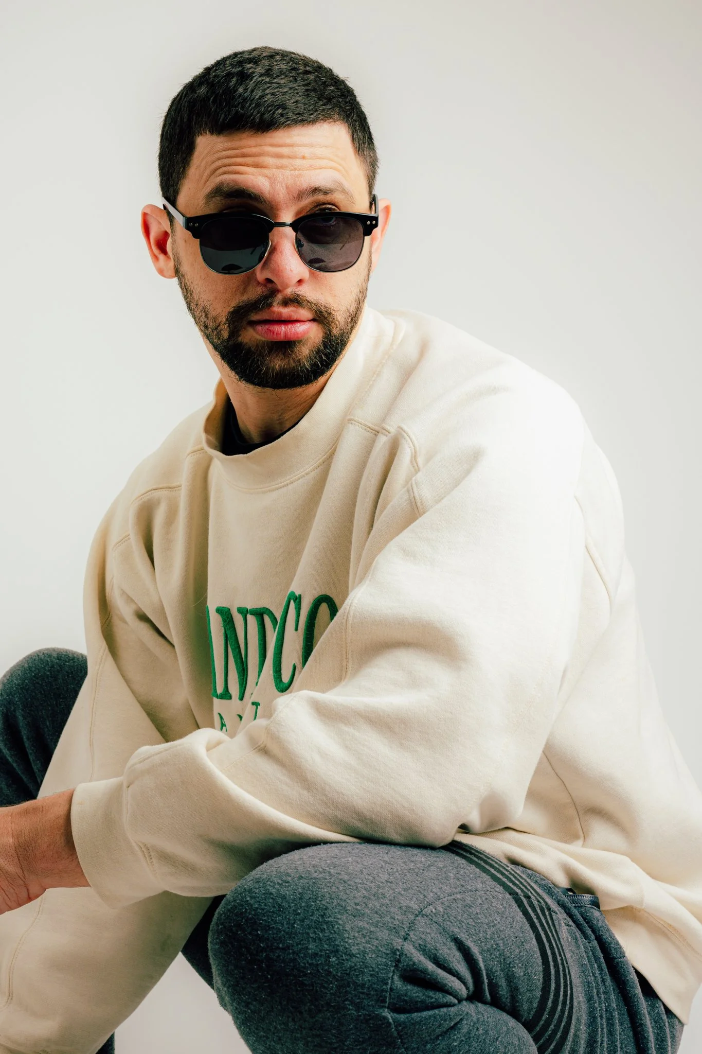A man with dark hair and a beard wearing black sunglasses and a cream-colored sweatshirt with green text, sitting against a plain background.