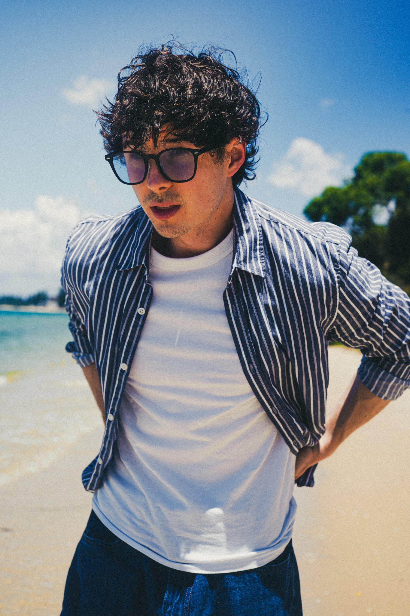 Young man with curly hair and sunglasses standing on a beach with a blue sky and water in the background.