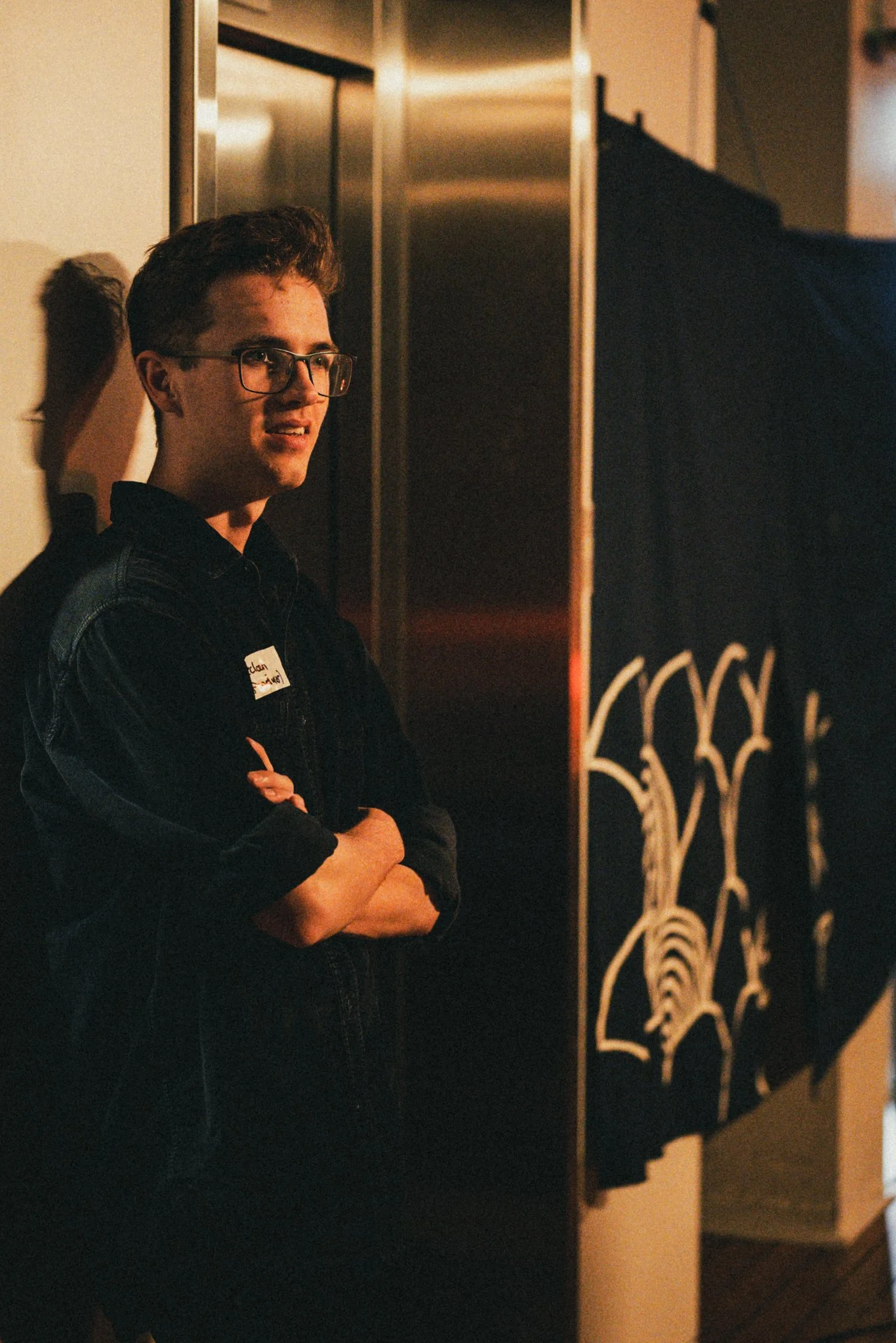 A young man with glasses, standing with arms crossed, in a dimly lit indoor setting.