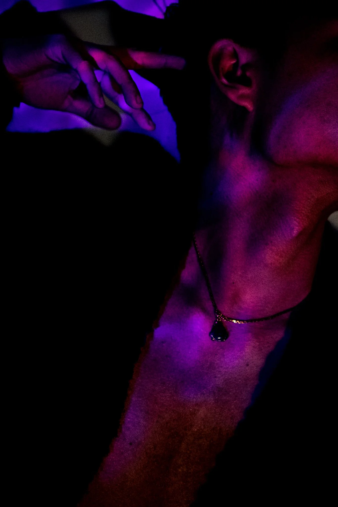 A person with short hair and a gold chain necklace with a dark pendant, in a dimly lit environment with purple and pink lighting, resting their head on their hand.