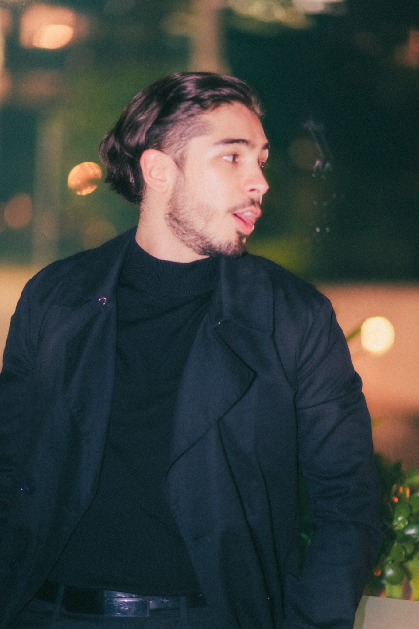 A young man with dark, wavy hair and a beard, wearing a black turtleneck and blazer, posing indoors at night with blurred city lights in the background.