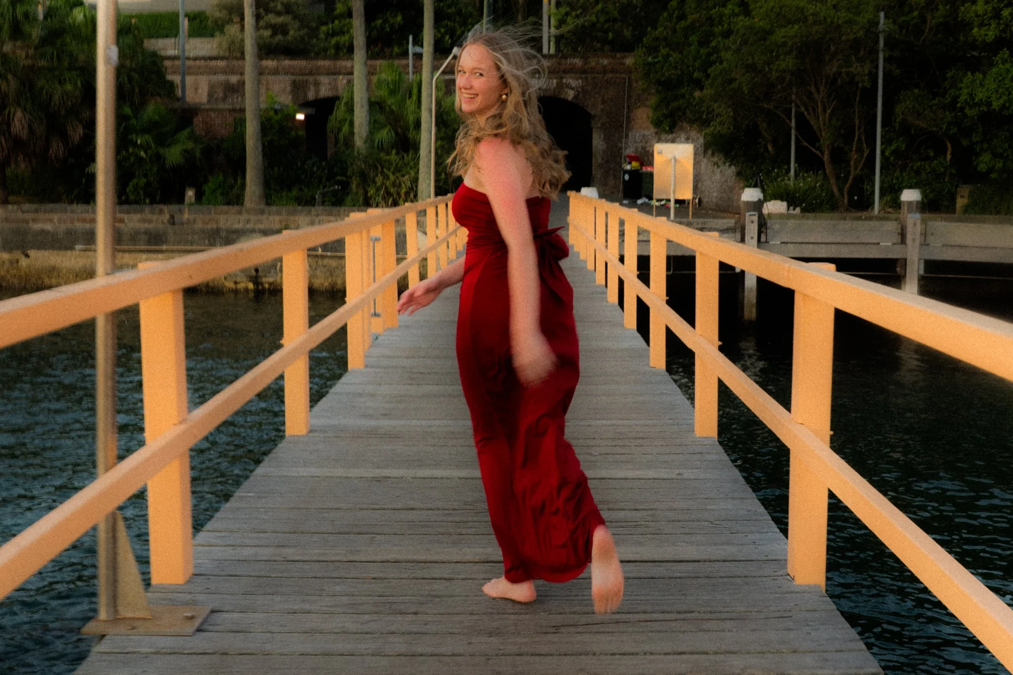 A woman in a red strapless dress walking barefoot on a wooden dock over water, smiling and looking back at the camera during sunset.