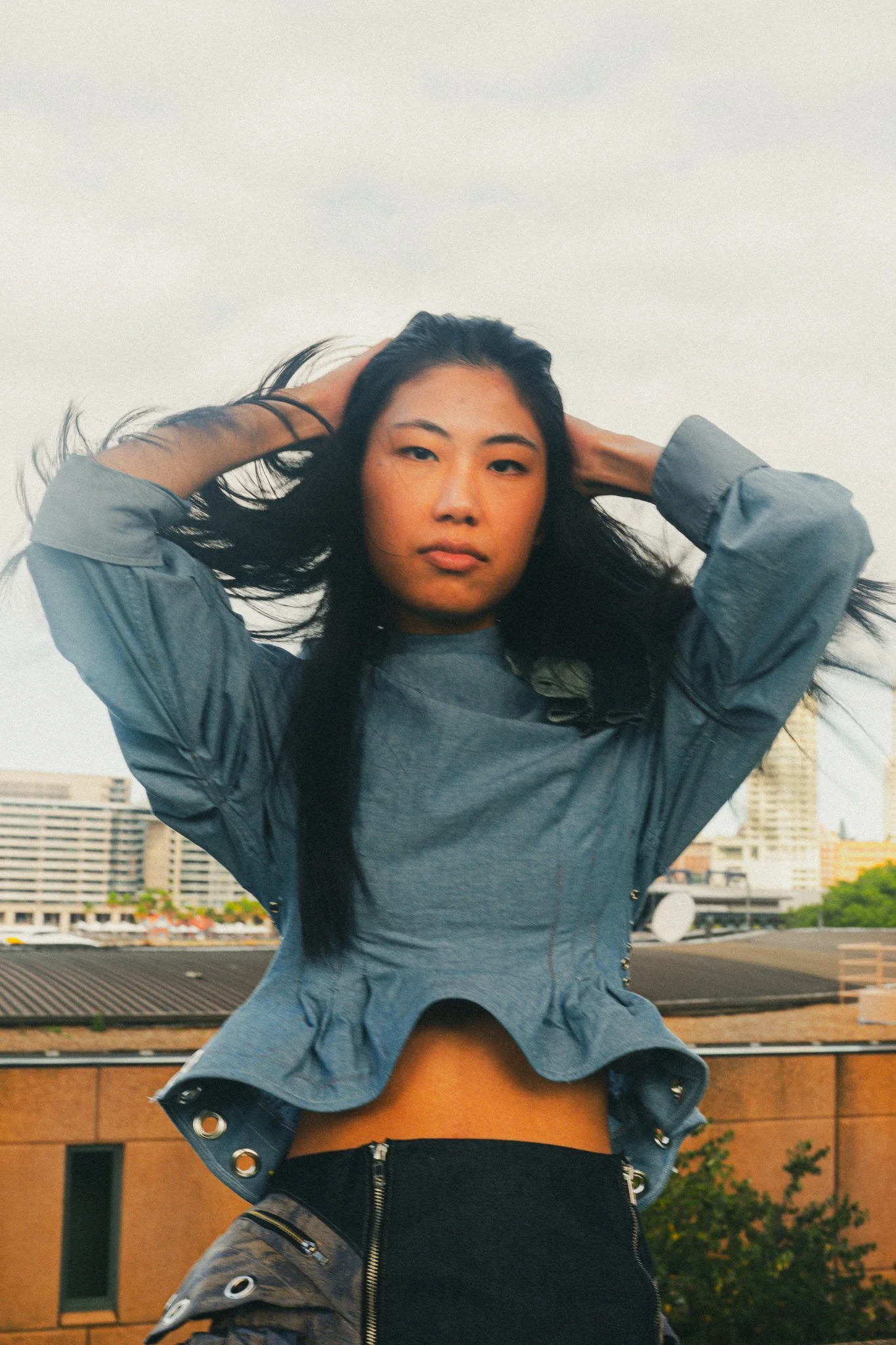 Young woman standing outdoors with her hands behind her head, wearing a gray jacket and black pants, cityscape in the background.